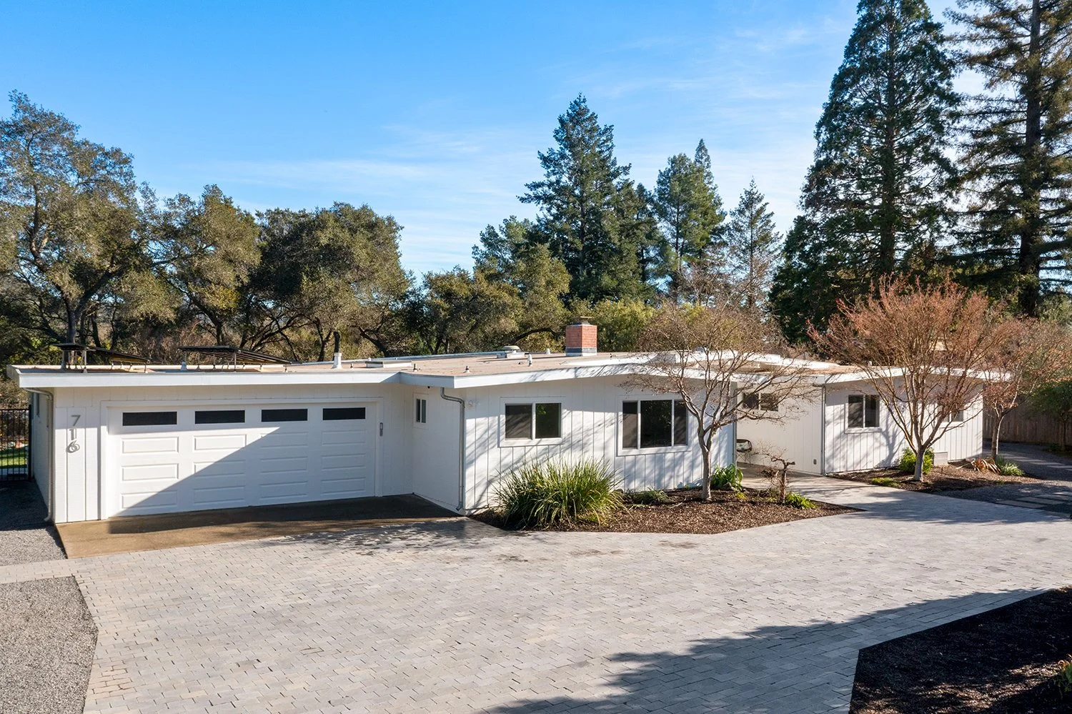 A modern single-story white house with a two-car garage, surrounded by trees and a paved driveway, under a blue sky.