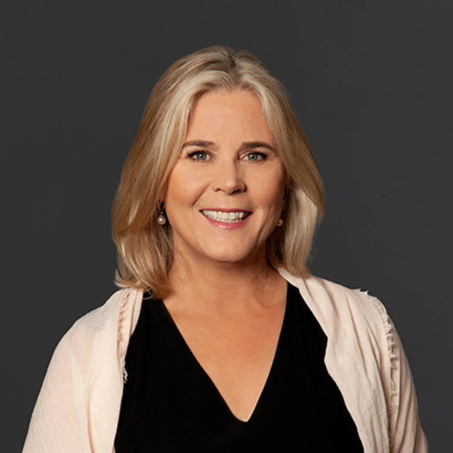 A professional headshot of a smiling blonde woman wearing pearl earrings, a black top, and a beige blazer against a dark background.