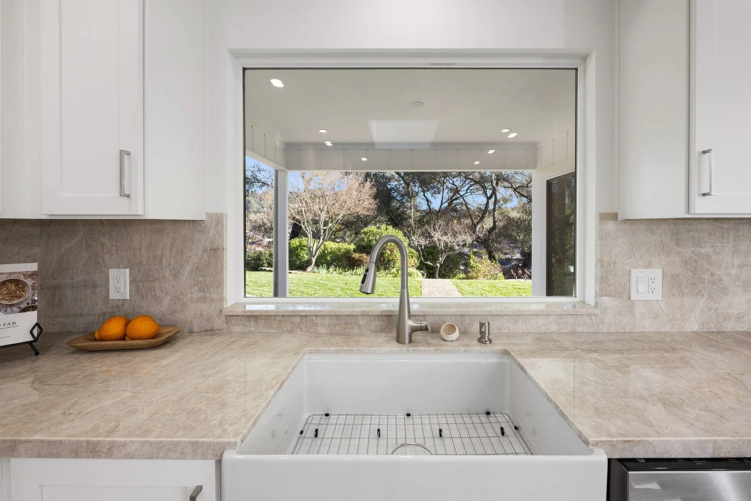 Kitchen sink area with a large window overlooking a garden with trees and bushes, beige countertop, white cabinets, three oranges on a tray, and electrical outlets.