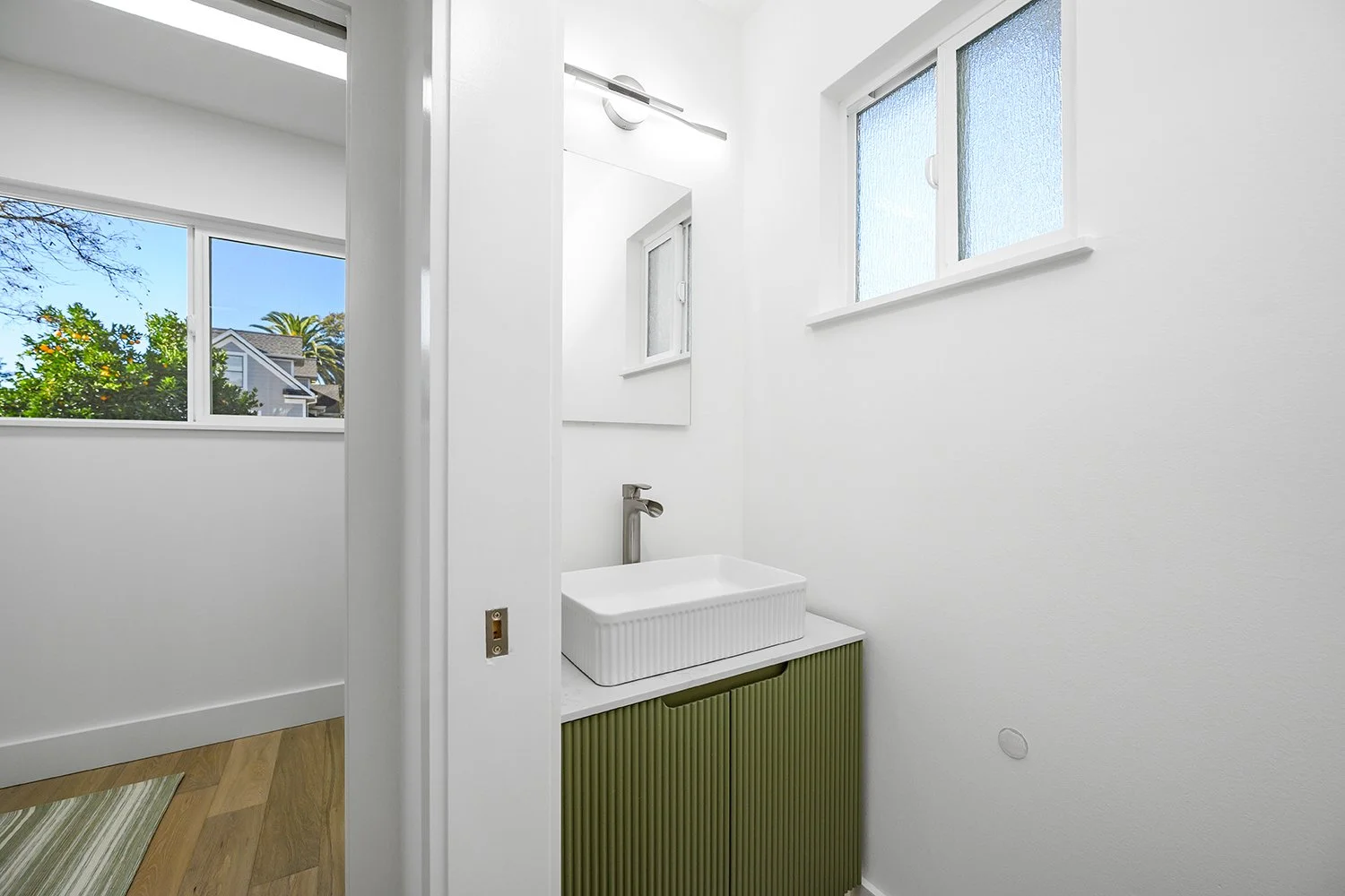 Small bathroom with a white rectangular sink on a green ribbed cabinet, above a window with frosted glass, and a larger window showing trees outside.
