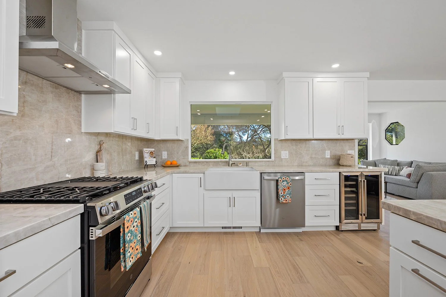 Modern kitchen with white cabinets, beige countertops, stainless steel appliances including oven and dishwasher, wood flooring, window above sink showing trees outside, and a view of a living area with a gray sofa.