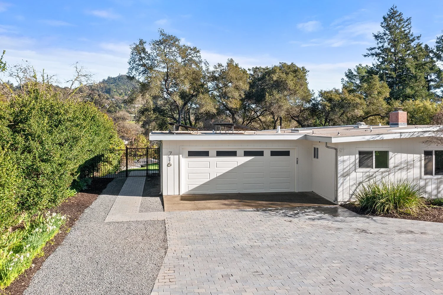 Front view of a modern white house with an attached garage, brick driveway, landscaped yard, and trees in the background under a blue sky.
