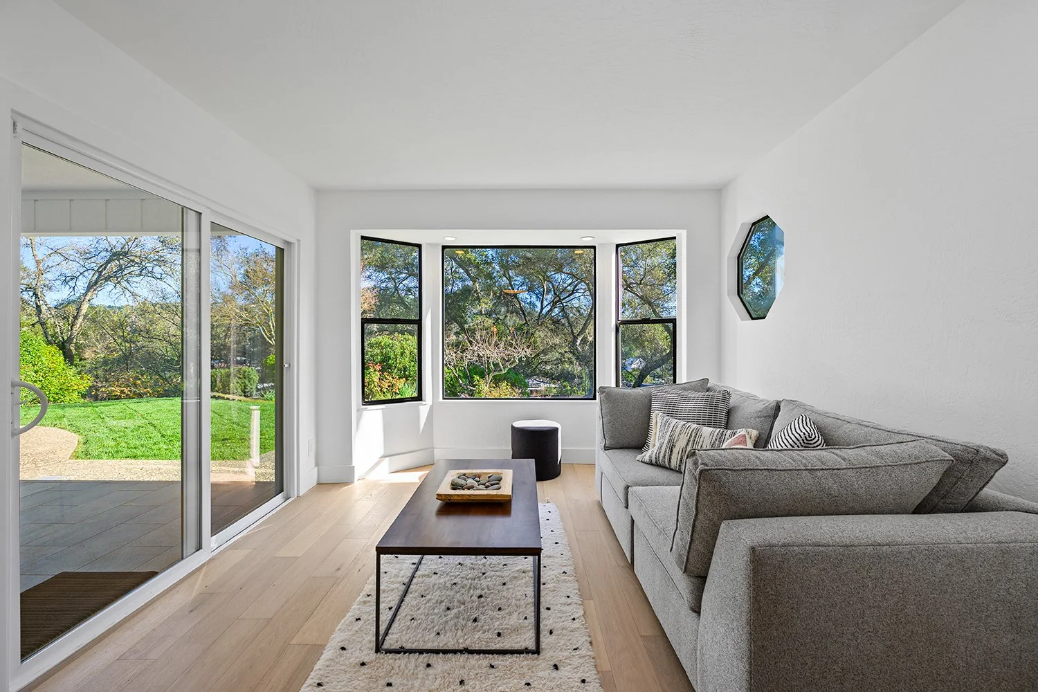 Living room with large bay window, gray sofa with pillows, wooden coffee table on a patterned rug, sliding glass door to patio, outside greenery and trees.
