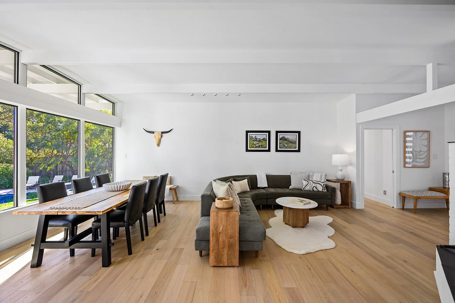 Open-concept living and dining room with wooden floors, large windows, a gray sectional sofa, a wooden dining table with black chairs, and wall art.