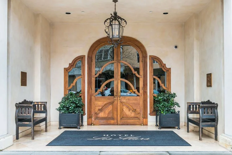 Elegant hotel entrance with wooden double doors, arched windows, potted plants, black benches, a hanging lantern, and a welcome mat.