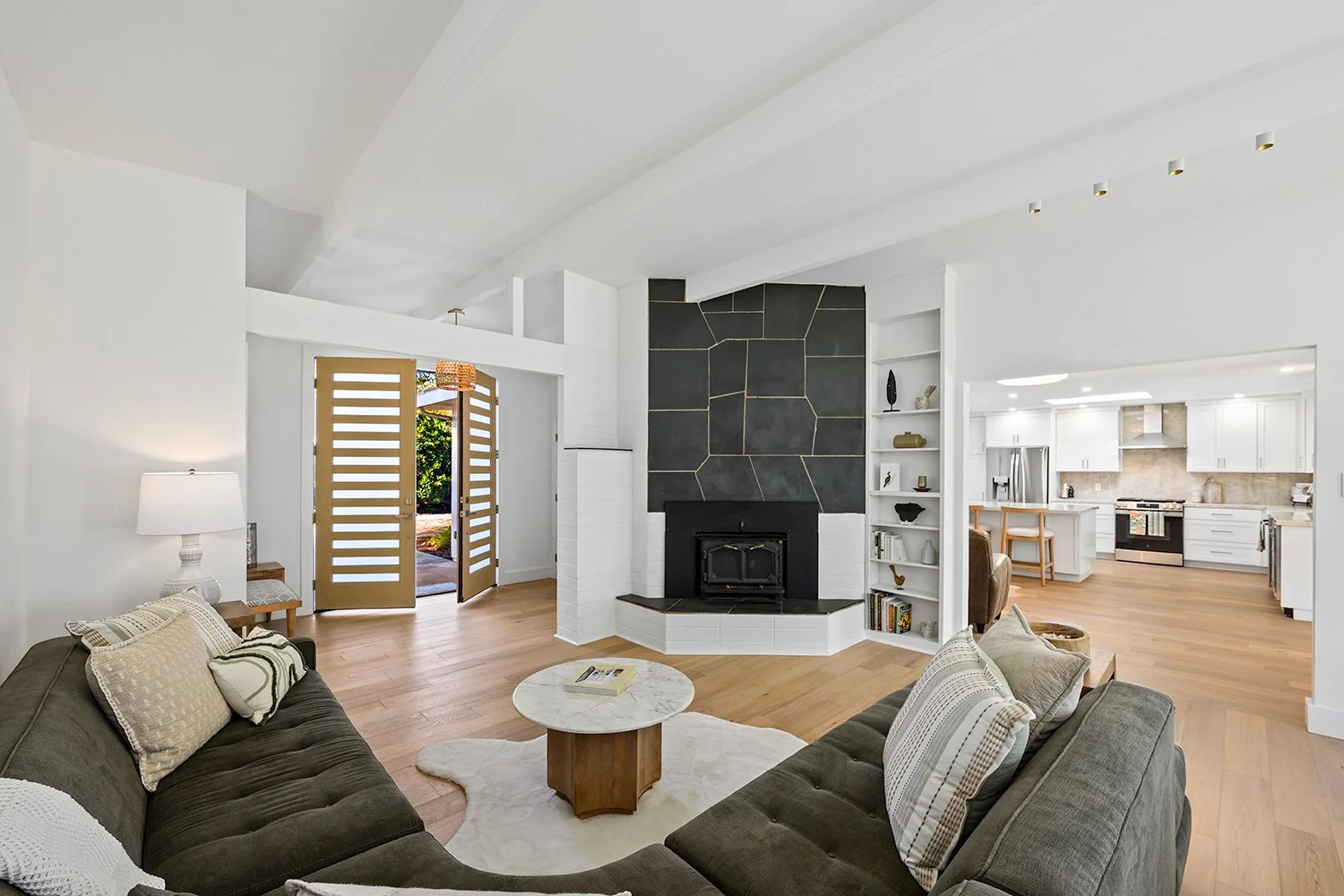 Living room with dark gray sectional sofa, white walls, and a fireplace with a black and gray geometric tile design, open to a kitchen with white cabinets and stainless steel appliances.