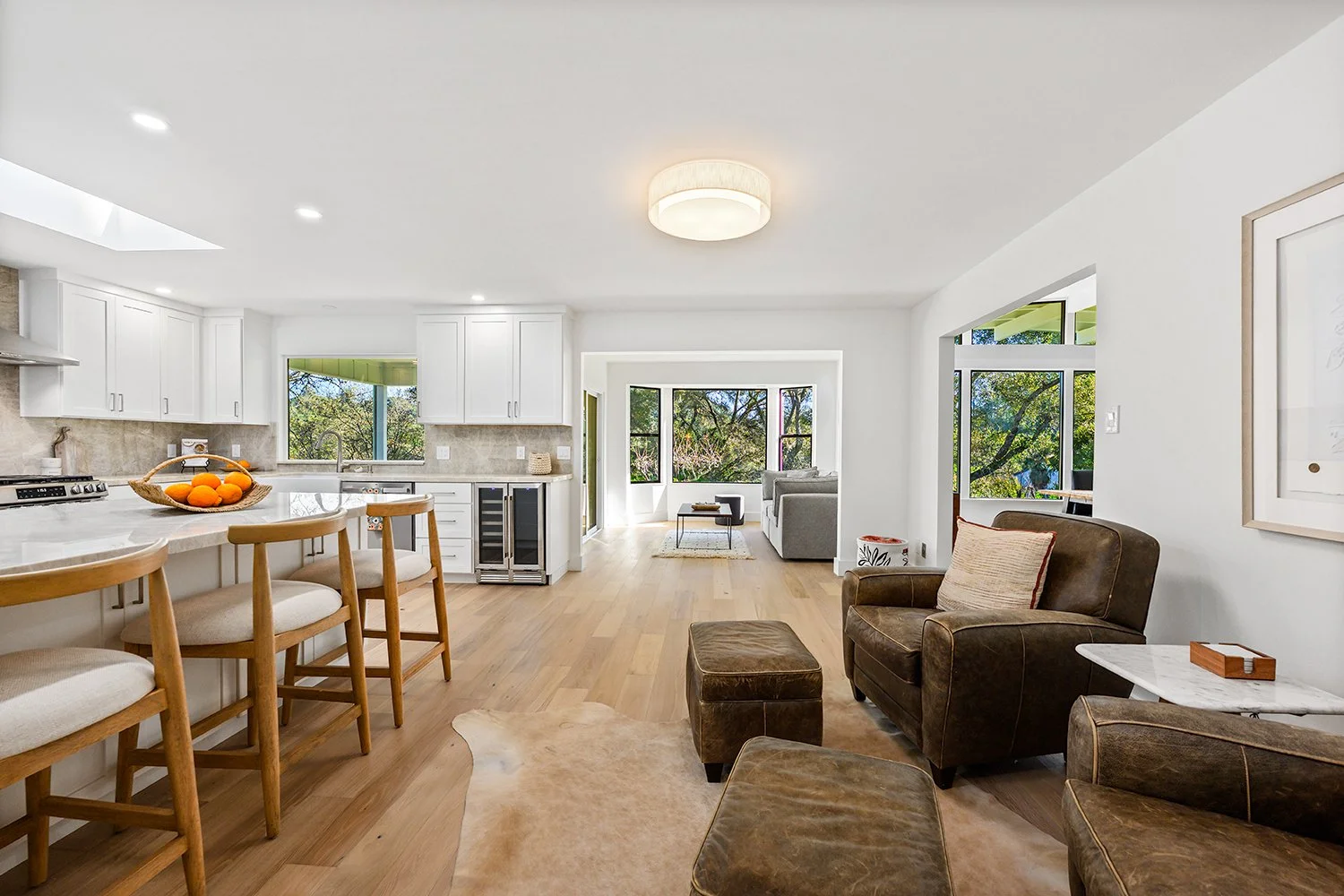 Open-concept living room and kitchen with white cabinets, wooden bar stools, leather chairs, and large windows showing trees outside.
