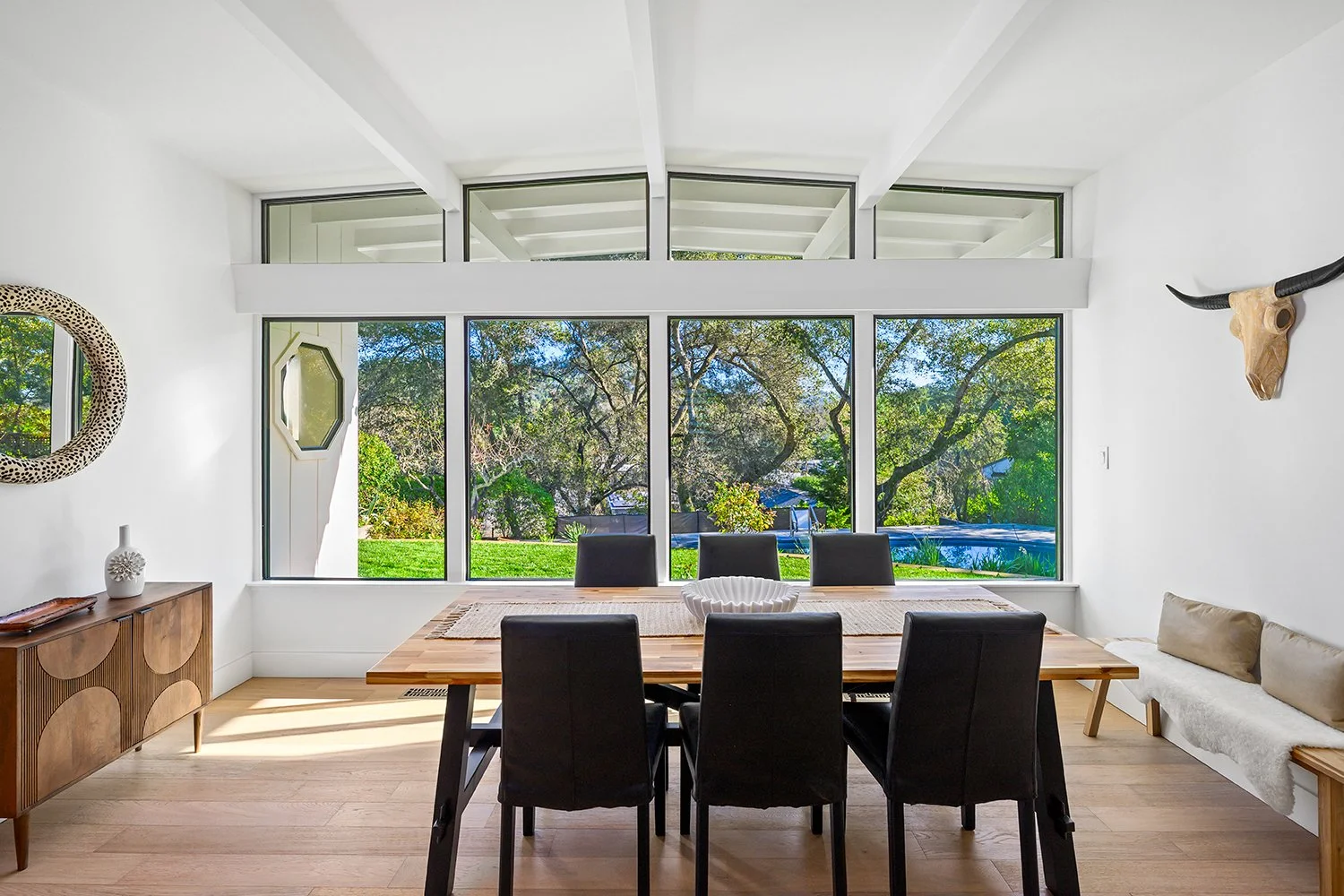 Bright dining room with large front window, wooden table, six black chairs, sideboard with decor, and wall decor including a giraffe skull with horns and a leopard print mirror.