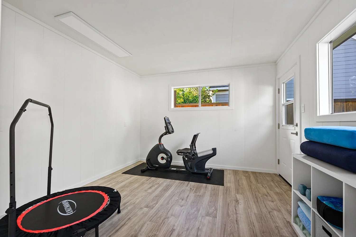 Room with wooden floor, white walls, a small window, and an exercise bike. There is a trampoline with a red border and black mat, and a shelving unit holding folded towels and weights.