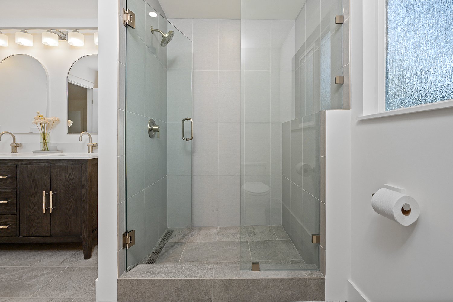 Modern bathroom with glass-enclosed shower, white tiled walls, gray floor tiles, wooden vanity with two faucets, mirror, and frosted window.