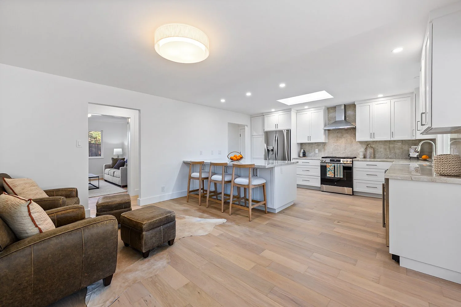 Open kitchen with white cabinets, stainless steel refrigerator, oven, and range hood, light wood flooring, bar stools at island, living room with brown leather sofa, and doorway to another room with a couch and window.