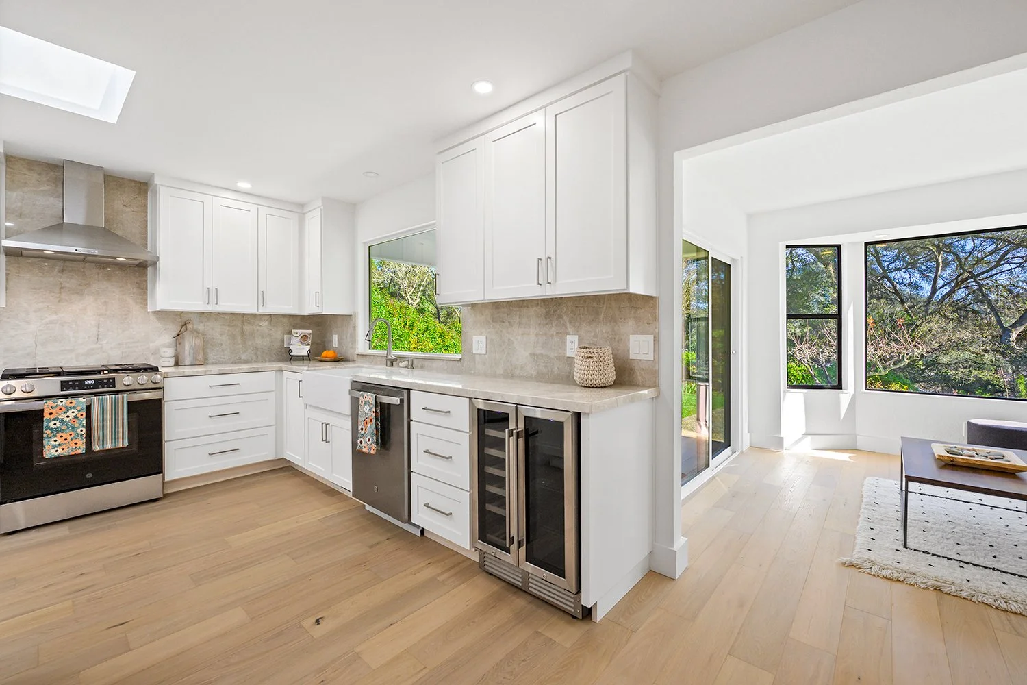 Modern kitchen with white cabinetry, beige countertops, stainless steel appliances, wood flooring, large windows showing outdoor greenery, and a bright adjoining living area.