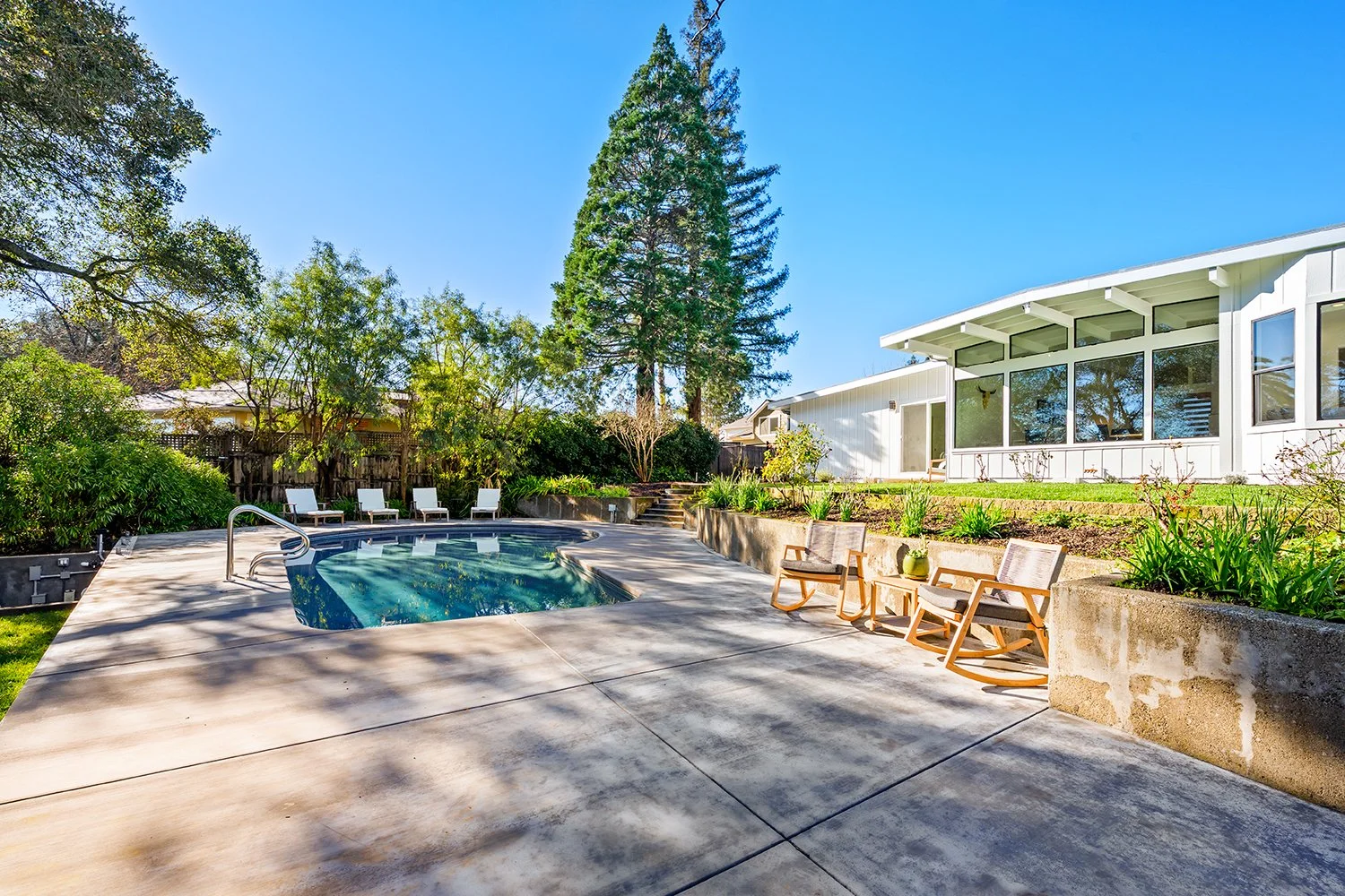 Backyard patio with a small swimming pool, outdoor chairs, and lush greenery under a clear blue sky.