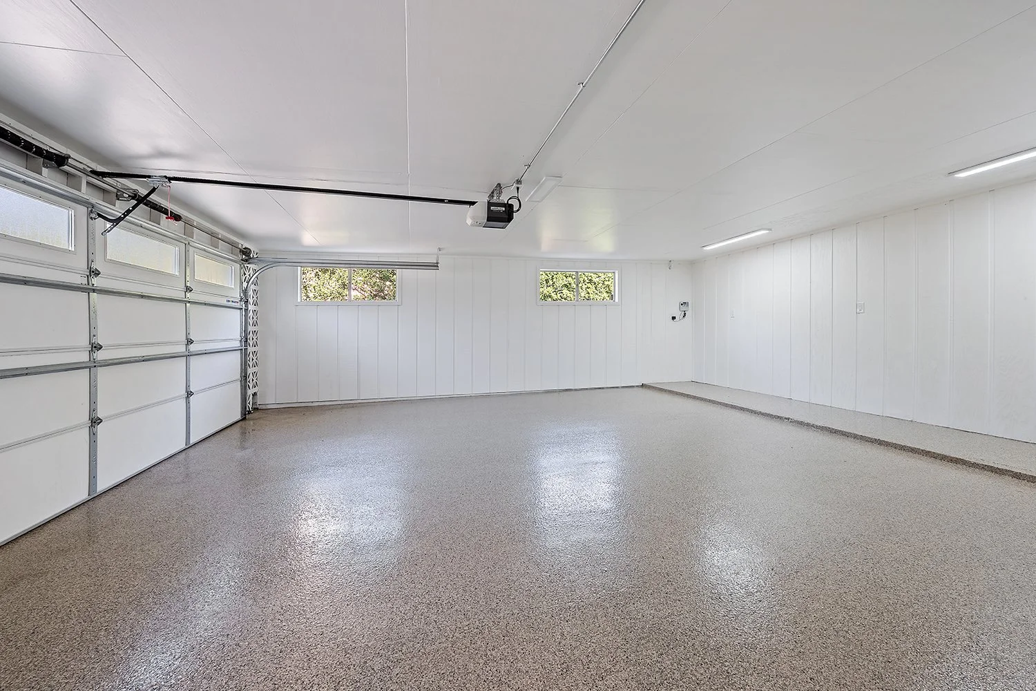 Empty garage with white walls, a closed white garage door with windows, small windows with greenery outside, and a polished concrete floor.