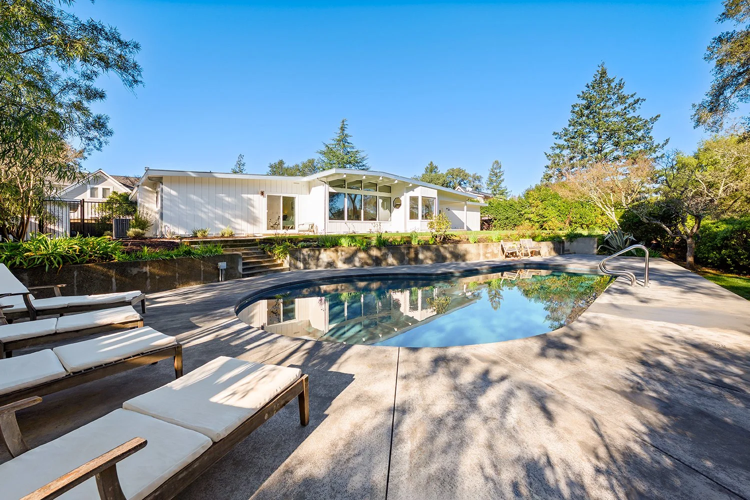 A backyard swimming pool with four lounge chairs, lush trees, and a modern white house in the background during daytime.