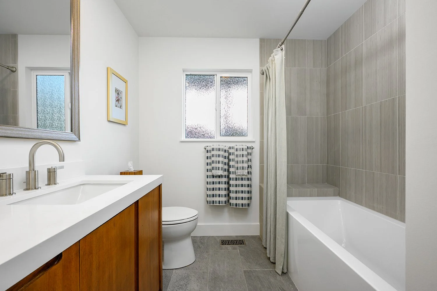 A clean, modern bathroom with a white sink and wooden vanity, a mirror, a toilet, and a bathtub with a shower curtain. There is a window with frosted glass, a framed picture on the wall, and striped towels hanging on a rack.