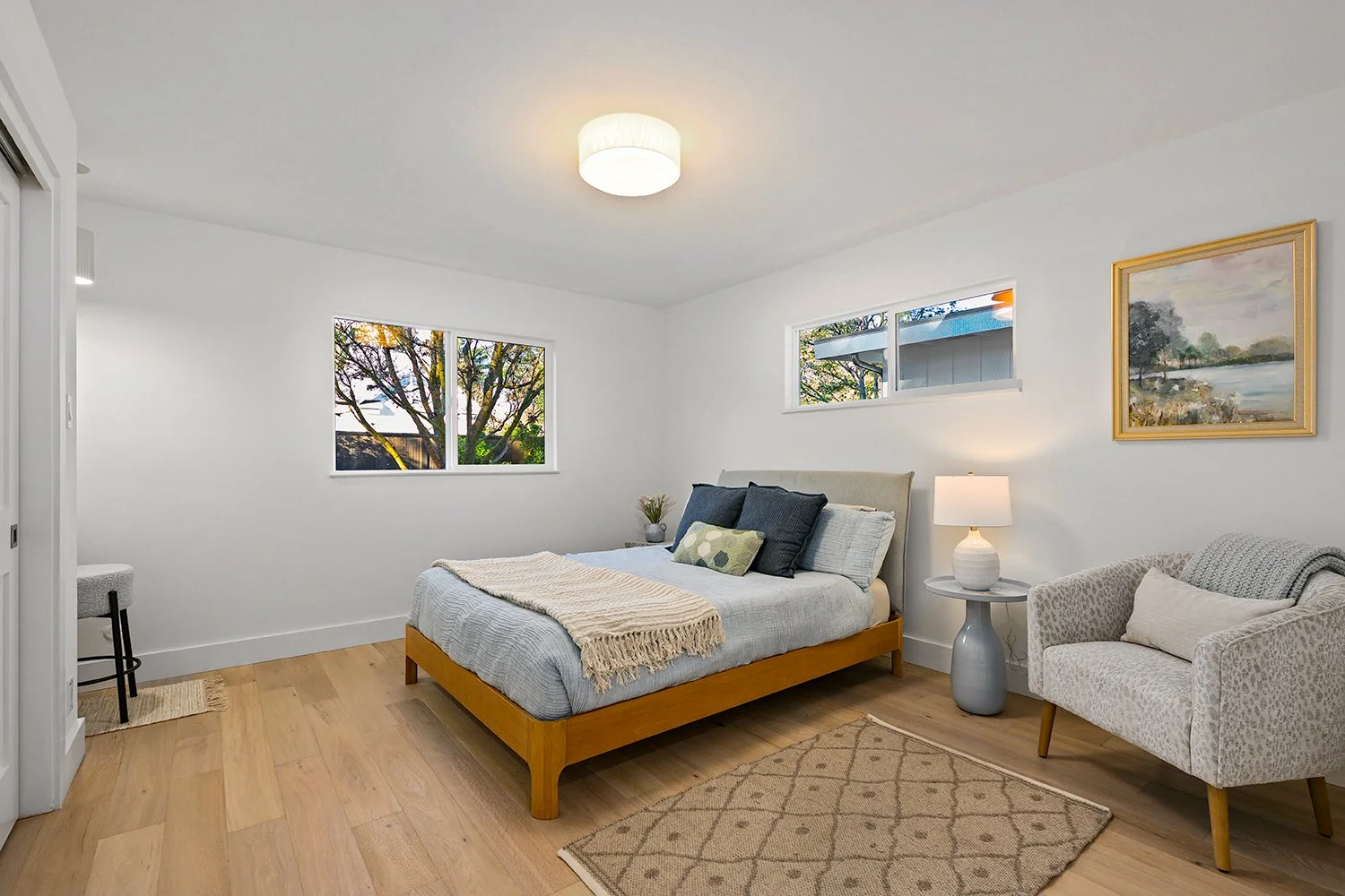 A bedroom with white walls, a wooden bed frame with gray bedding, a beige armchair, a small round side table with a lamp, two windows showing trees outside, and a framed landscape painting on the wall.
