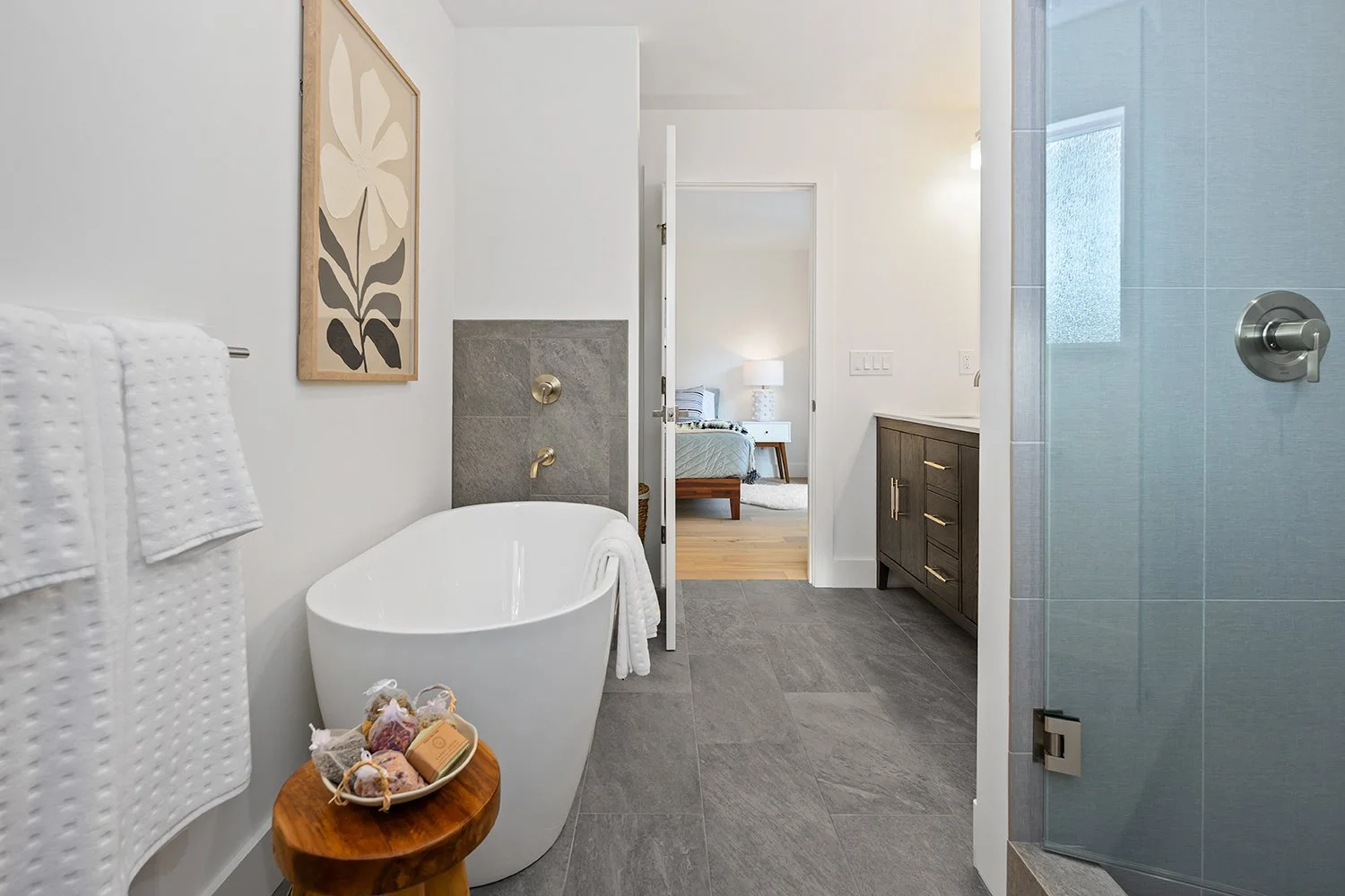 Modern bathroom with a white bathtub, gray tiled floor, and shower enclosure. A wooden stool with bath products is next to the tub. There is a framed artwork of a flower on the wall. An open door reveals a bedroom with a bed, bedside table, lamp, and