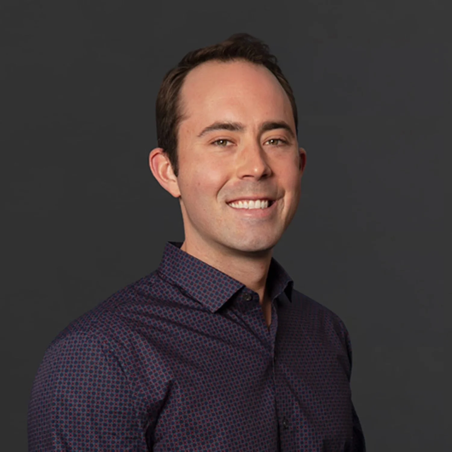 Headshot of a smiling young man with short dark brown hair, wearing a dark patterned button-up shirt against a plain dark background.
