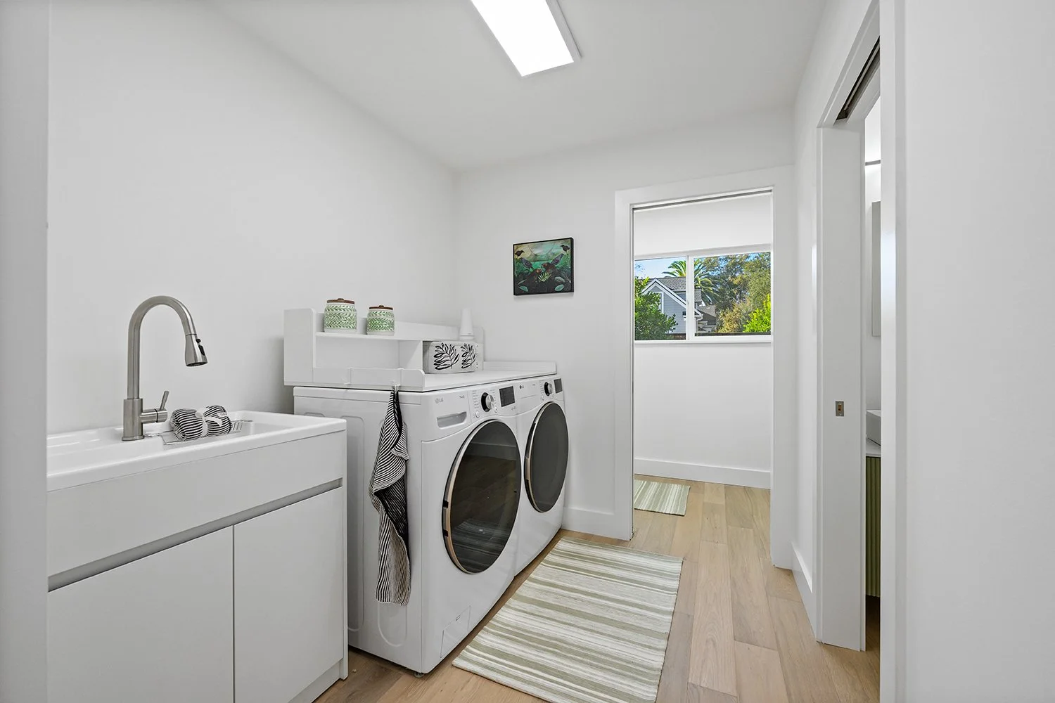 A bright laundry room with white walls, a sink with a faucet, a washing machine, a dryer, a window showing greenery outside, and a small picture hanging on the wall.