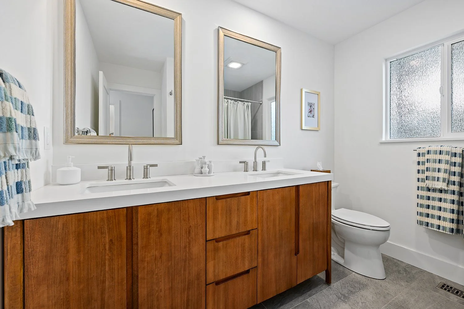Modern bathroom with double vanity, wooden cabinets, two mirrors, white countertops, toilet, and frosted window with striped towel.