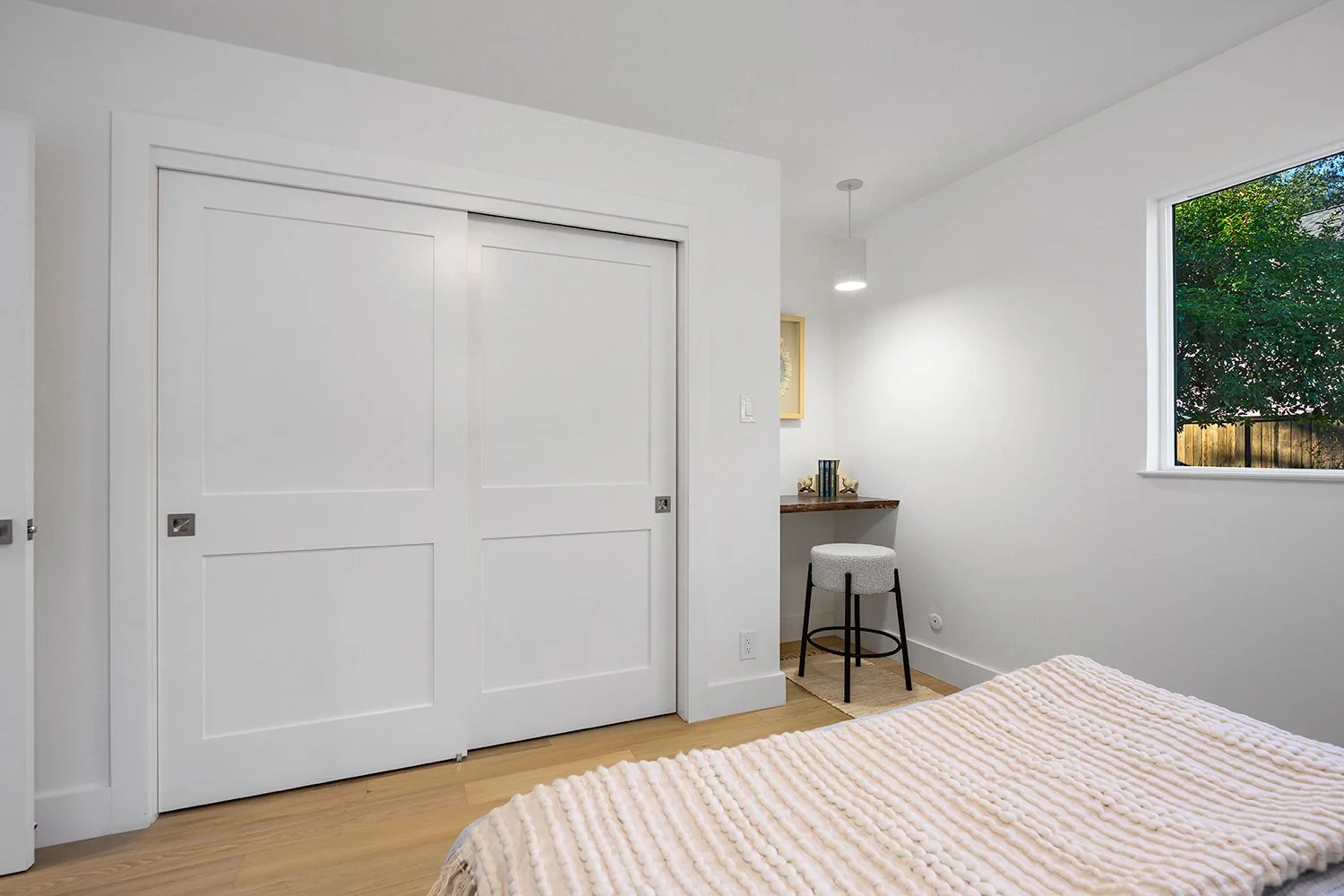 A minimalistic bedroom with white walls, a white closet with sliding doors, a window showing green trees outside, and a small workspace with a stool and a wooden shelf with books.
