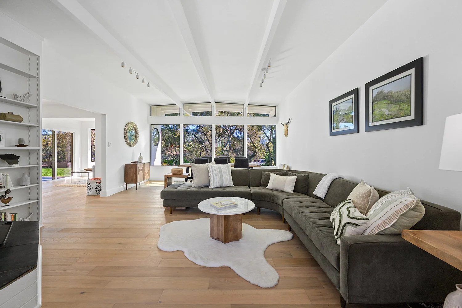Bright living room with large windows, gray sectional sofa with white and beige pillows, round marble-top coffee table on a white fluffy rug, wooden floor, and wall art.