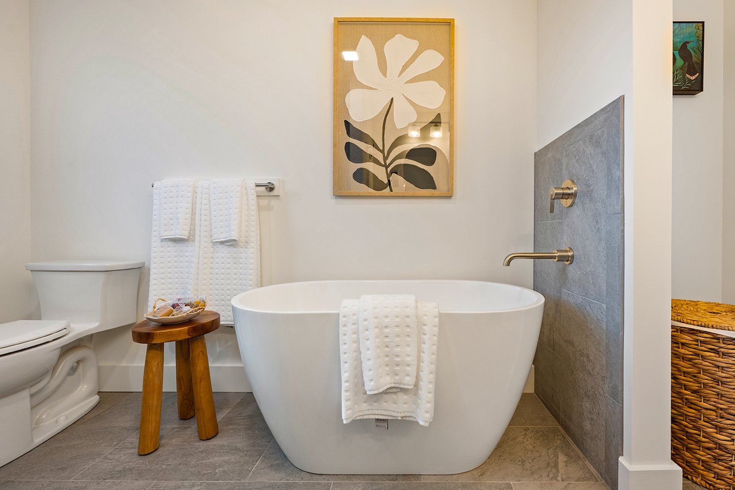 Modern bathroom with a free-standing bathtub, gray tiled wall, artwork of a large flower, towel rack, and a small wooden stool with toiletries.