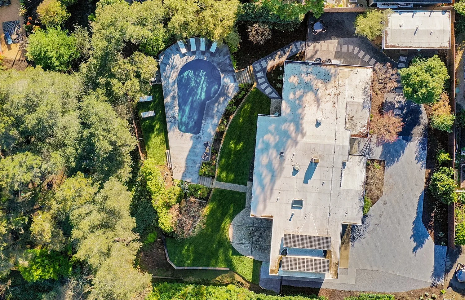 Aerial view of a house with a swimming pool, surrounding lush green trees, a lawn, sidewalks, and a parking area.