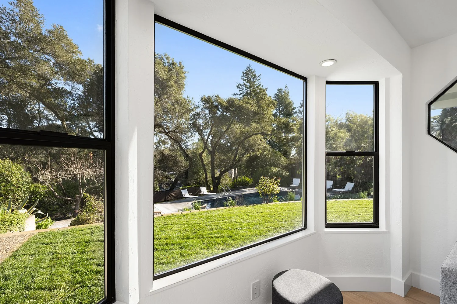 View from inside a bright, modern room with large black-framed windows overlooking a backyard with a swimming pool, green grass, trees, and outdoor lounge chairs.