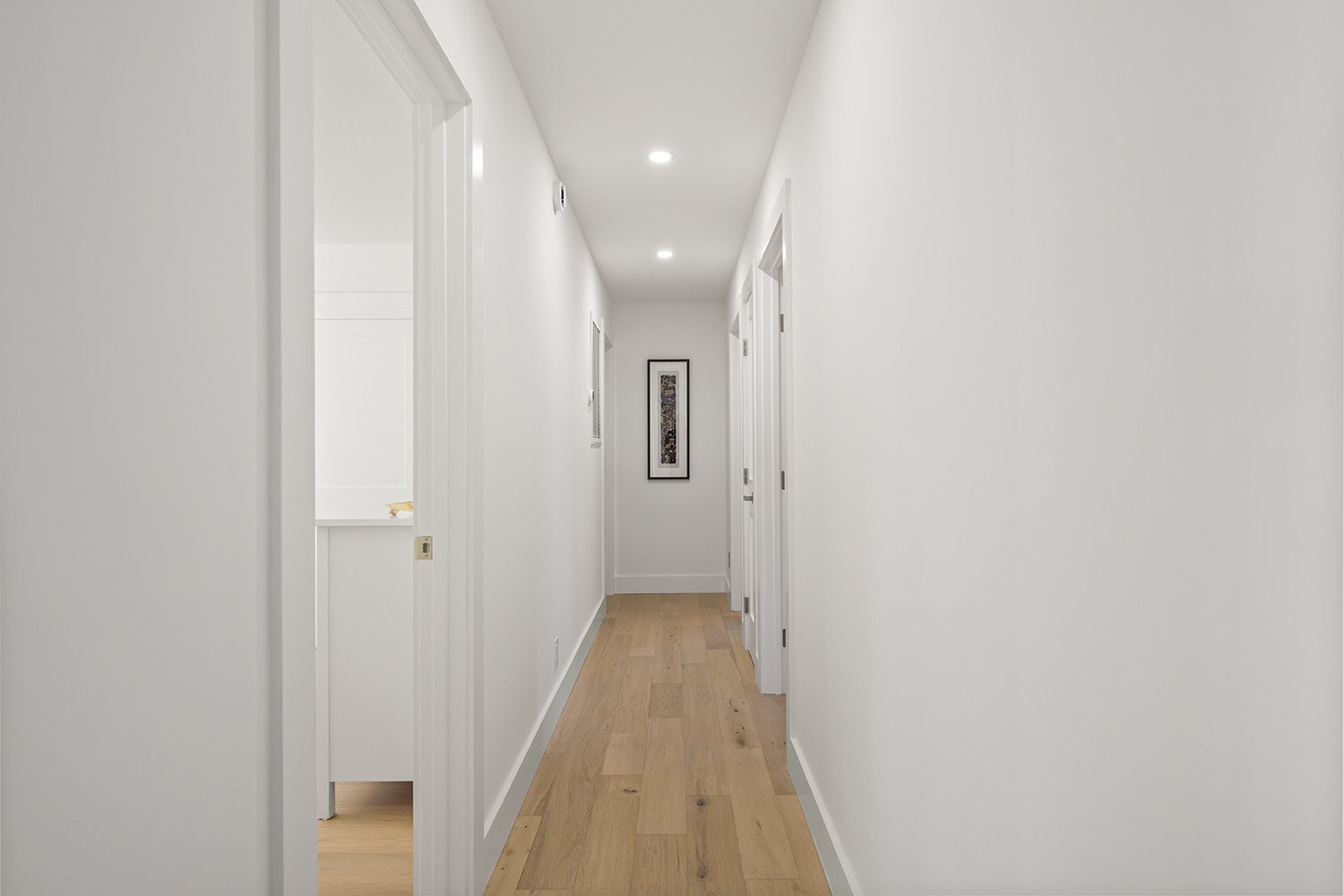A clean, narrow hallway in a modern home with white walls, wooden flooring, and recessed ceiling lights. A framed piece of art hangs on the far wall.