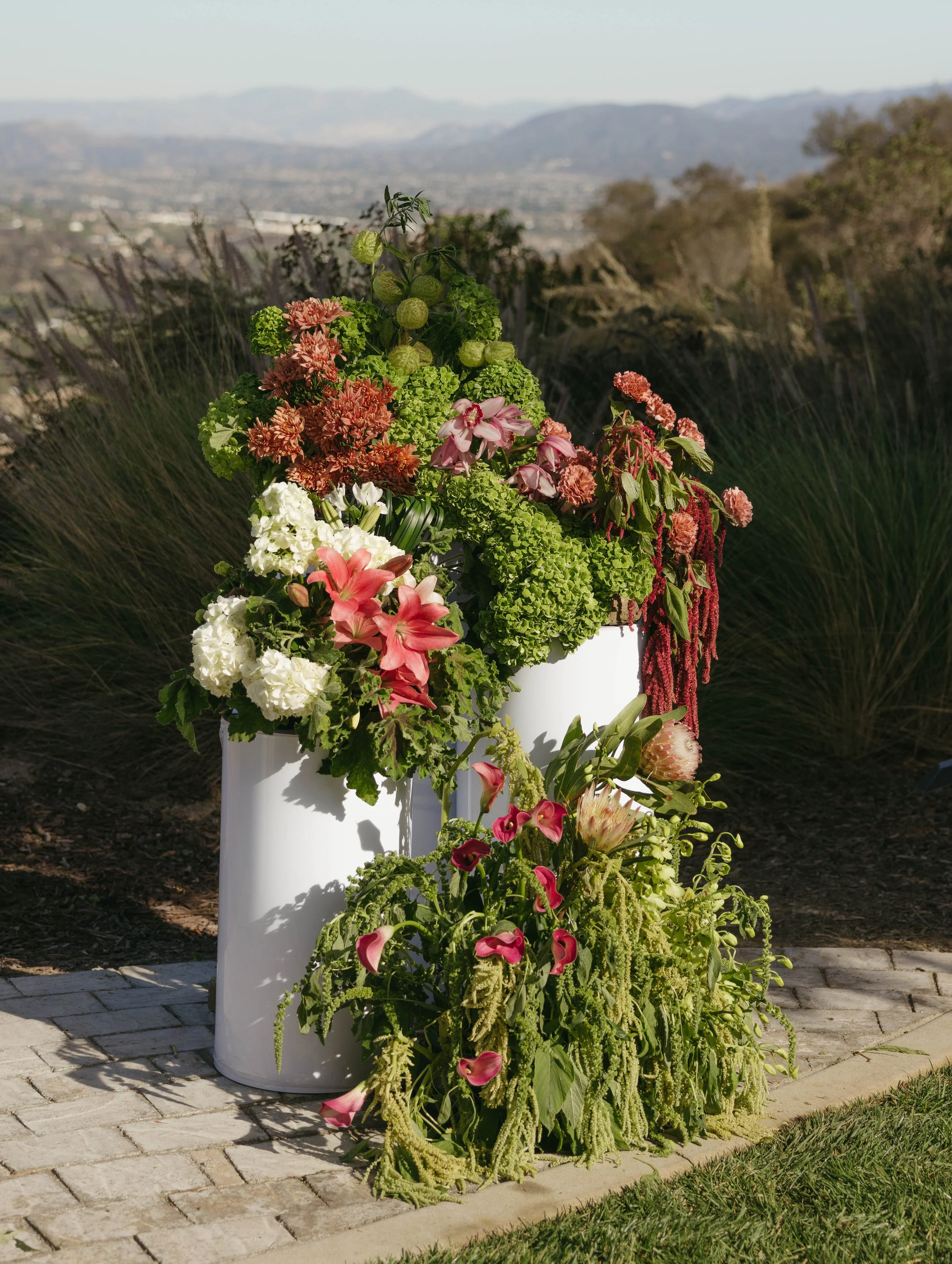 A large white planter with colorful flowers, including pink lilies, white hydrangeas, and other green and pink flowering plants, outdoors on a paved area with a mountainous landscape in the background.