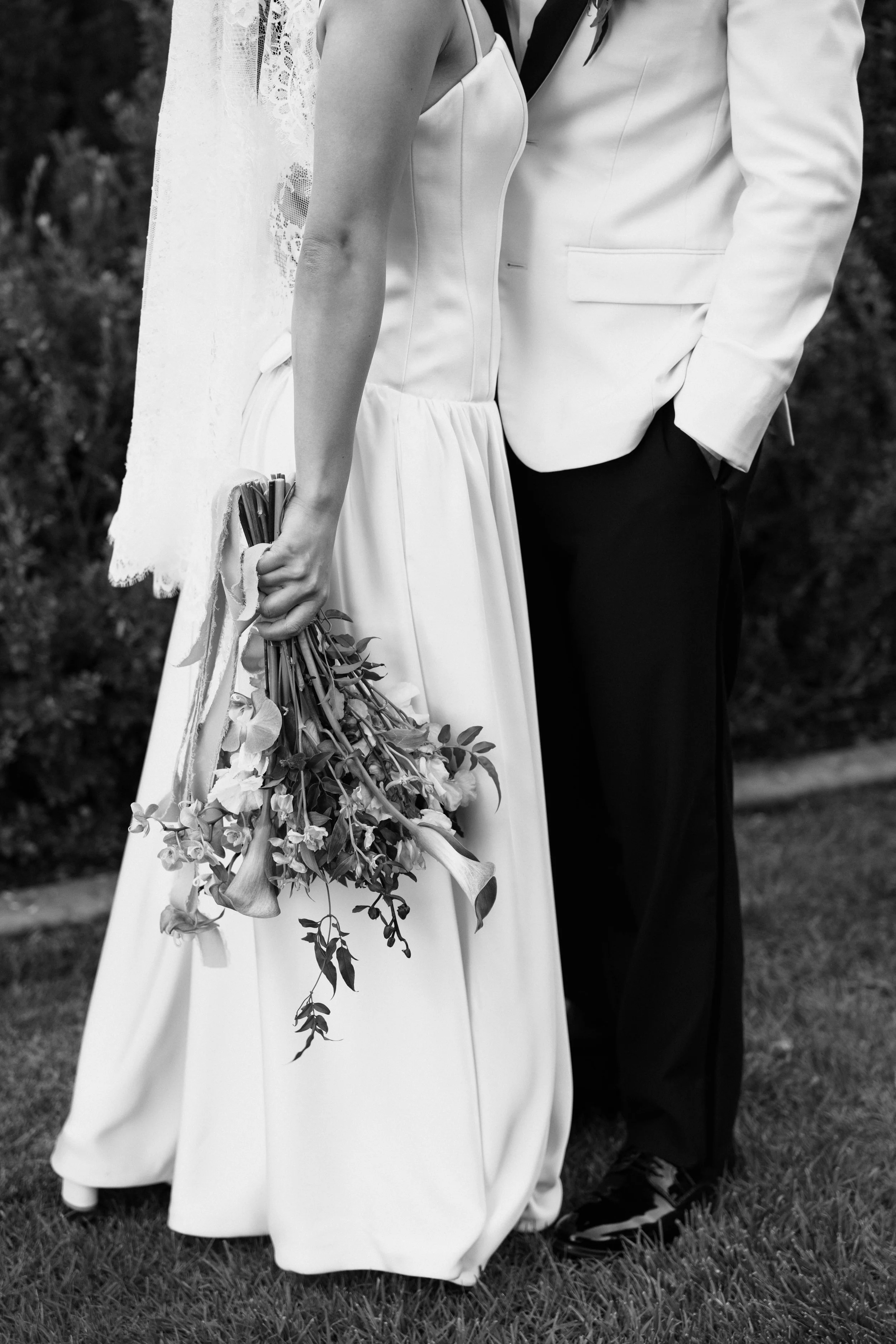 Black and white photo of a bride in a wedding gown holding a bouquet of flowers, standing close to a groom in a tuxedo, outdoors on grass.