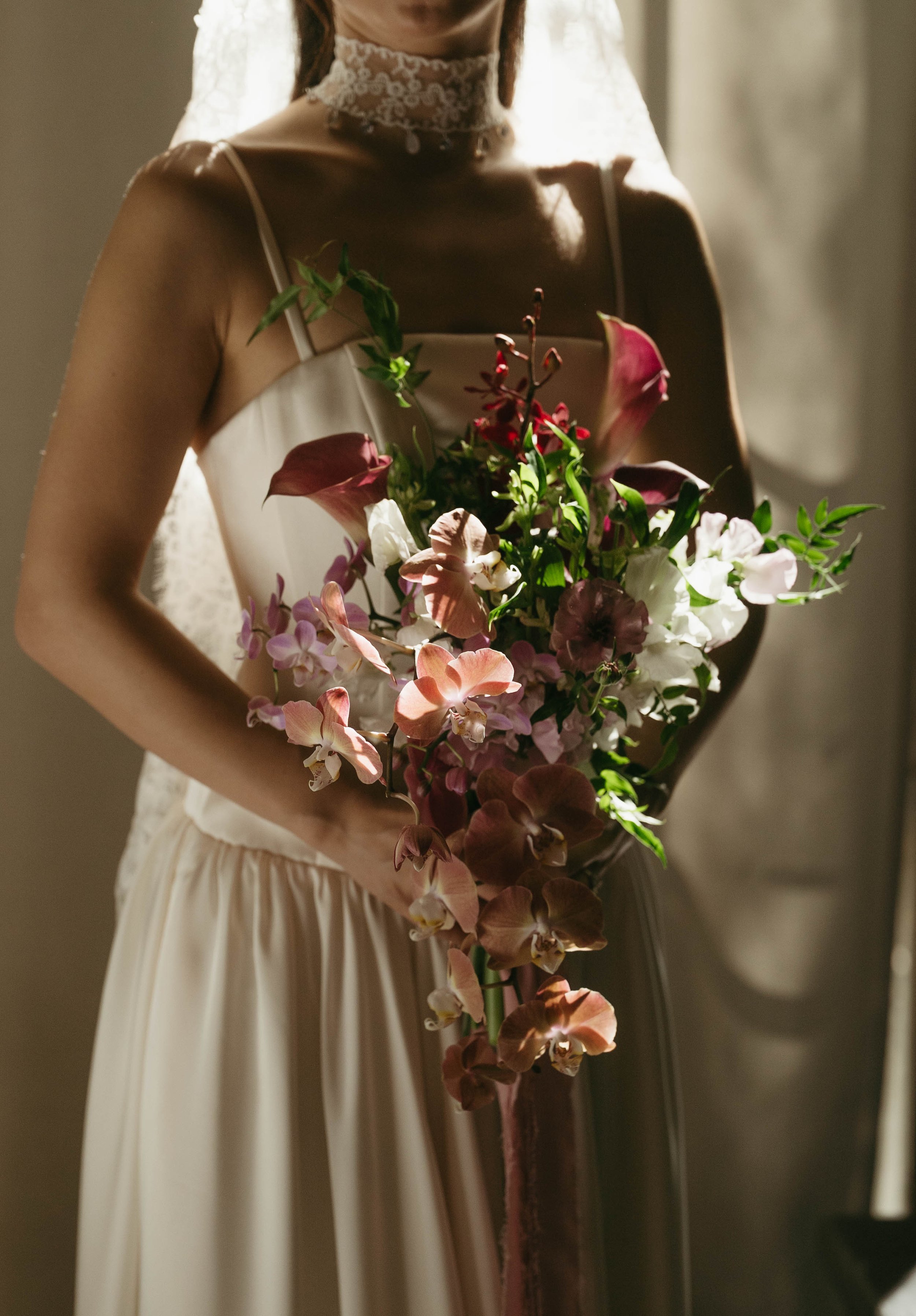 A woman dressed in a white dress with thin straps and lace at the neck, holding a large bouquet of mixed pink, white, and green flowers, standing in front of a neutral wall with soft lighting.