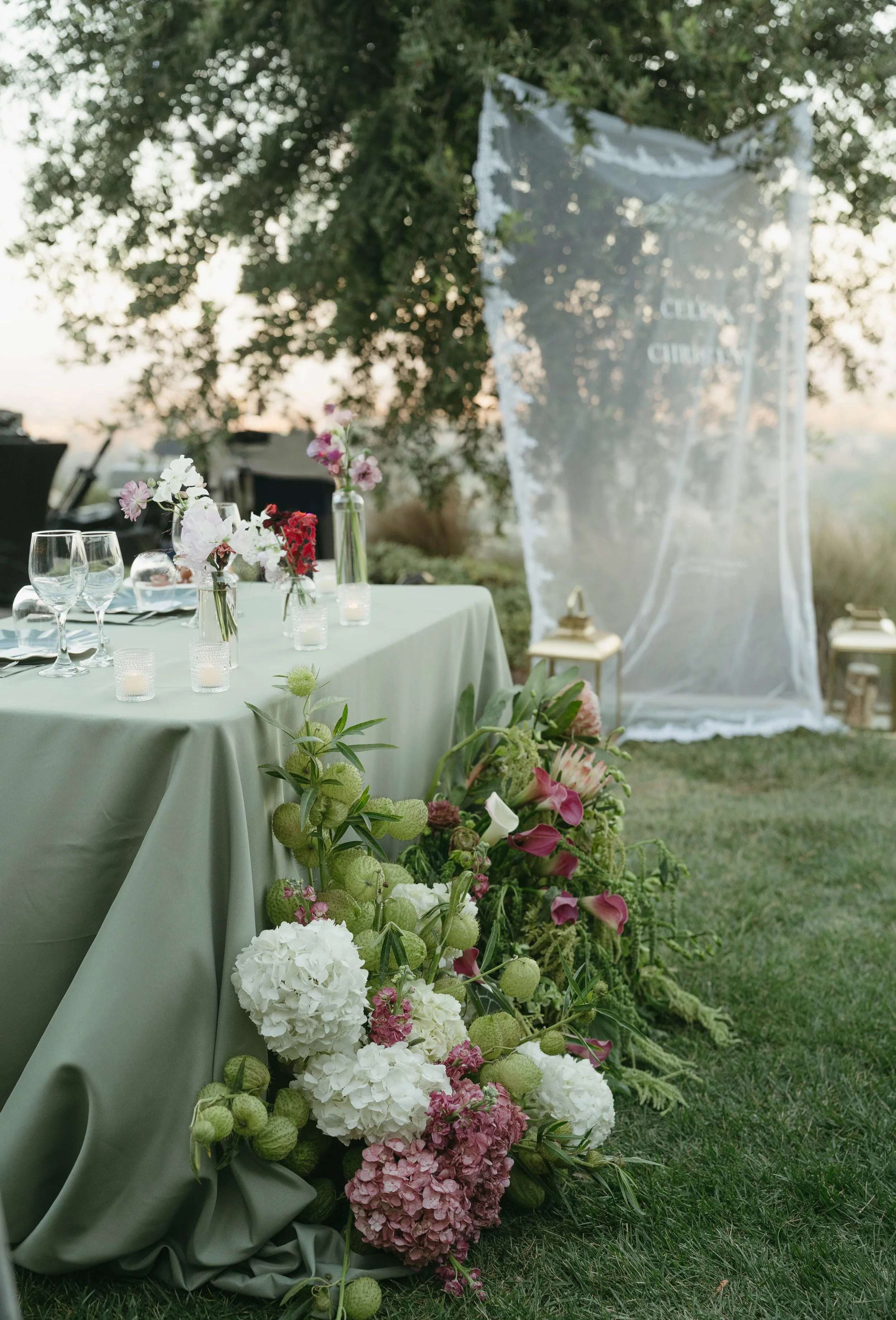 Outdoor event table decorated with floral arrangements and candles, with a sheer fabric backdrop and lanterns in the background