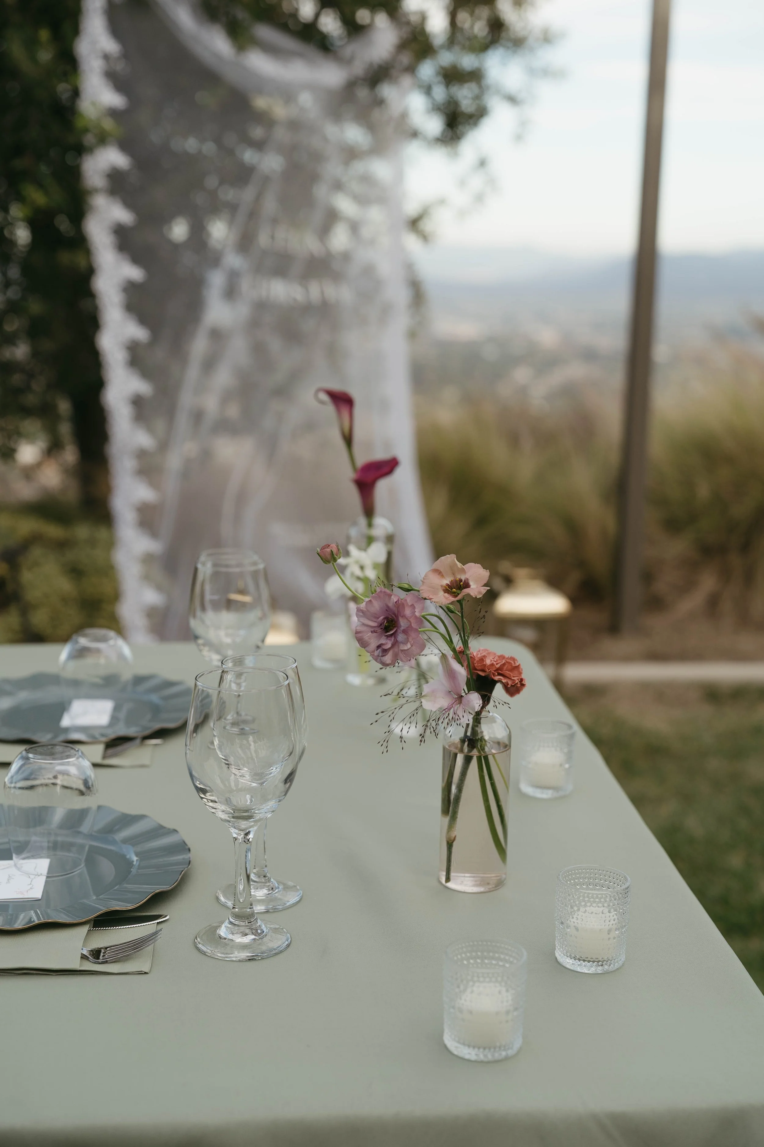 Elegant outdoor dining table with glassware, floral centerpiece, candles, and place settings on a pale green tablecloth, overlooking a scenic outdoor view.