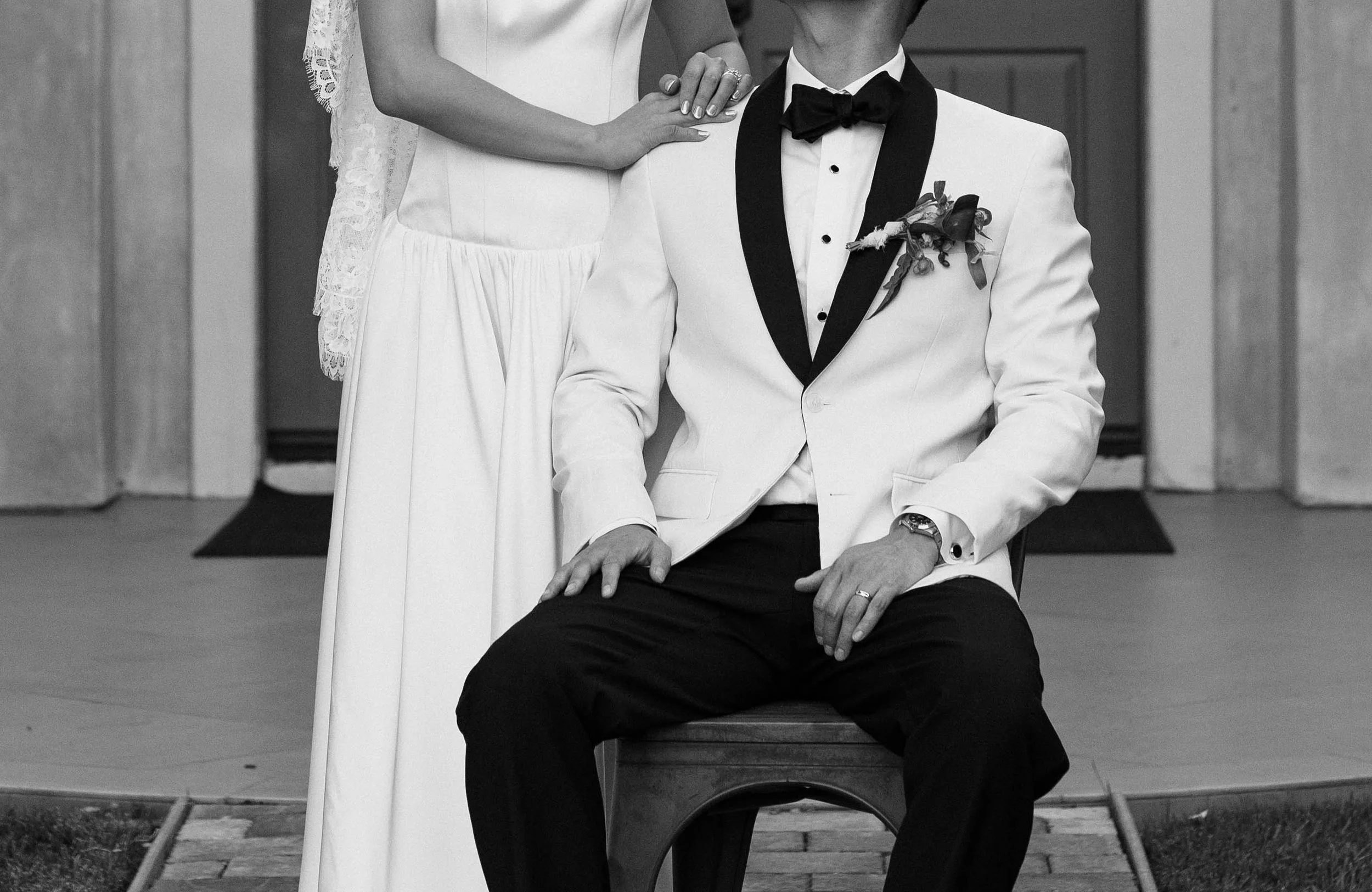 A black-and-white photo of a seated man in a tuxedo with a white jacket, black bow tie, and boutonniere, with a woman standing beside him, her hand resting on his shoulder, wearing a wedding dress with lace details.