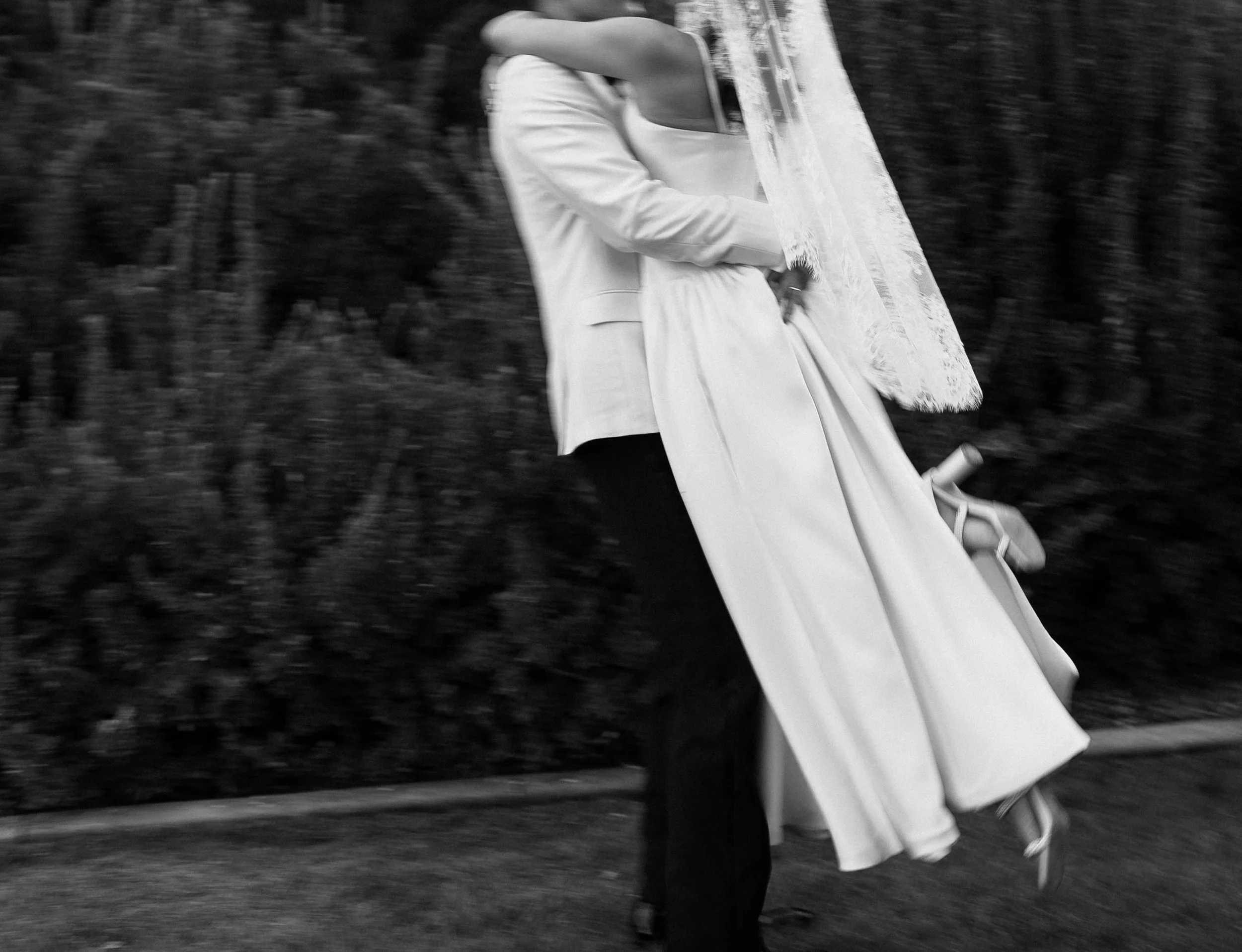 A black-and-white photo of a couple dancing, with the person in a wedding dress and the other in a suit, captured mid-movement outdoors.