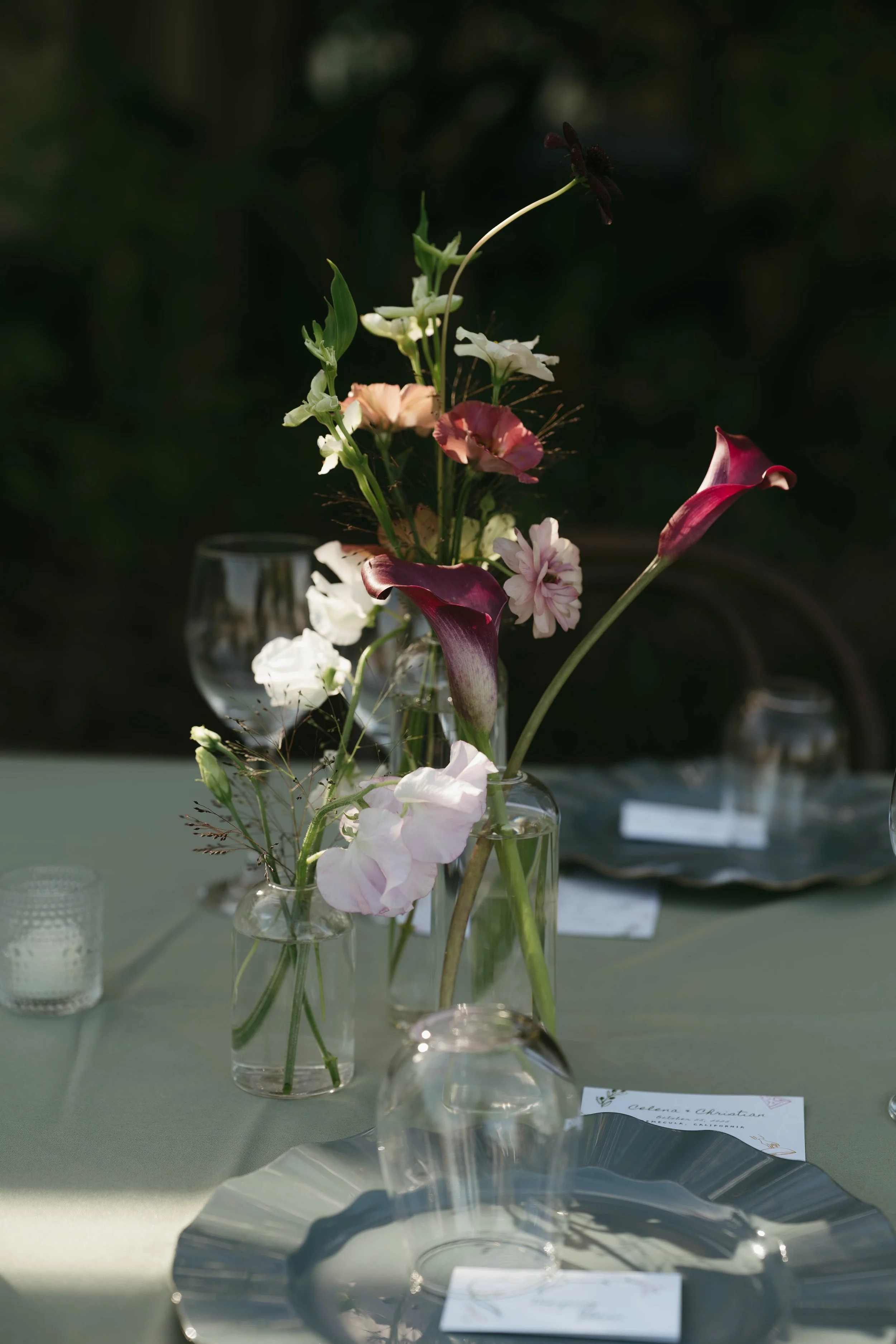 A table setting with a floral centerpiece in vases, including calla lilies and other flowers, with plates, glasses, and name cards.