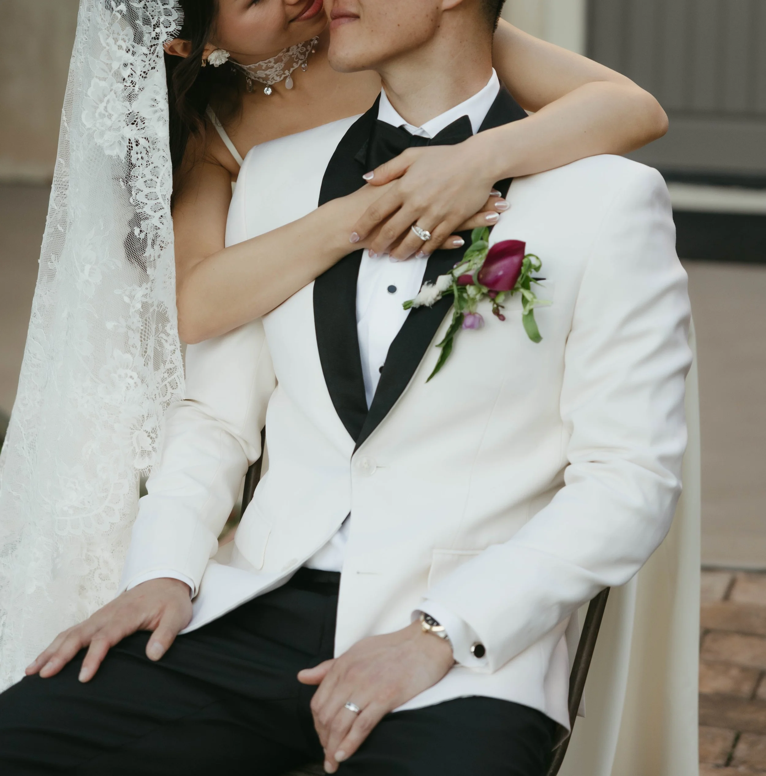 A bride and groom close-up during their wedding ceremony or celebration, with the bride leaning in towards the groom, his hand resting on his lap, and the bride's arms wrapped around his shoulders. The groom is wearing a white tuxedo jacket with black lapels and a boutonniere, and the bride is in a lace wedding dress with a veil.
