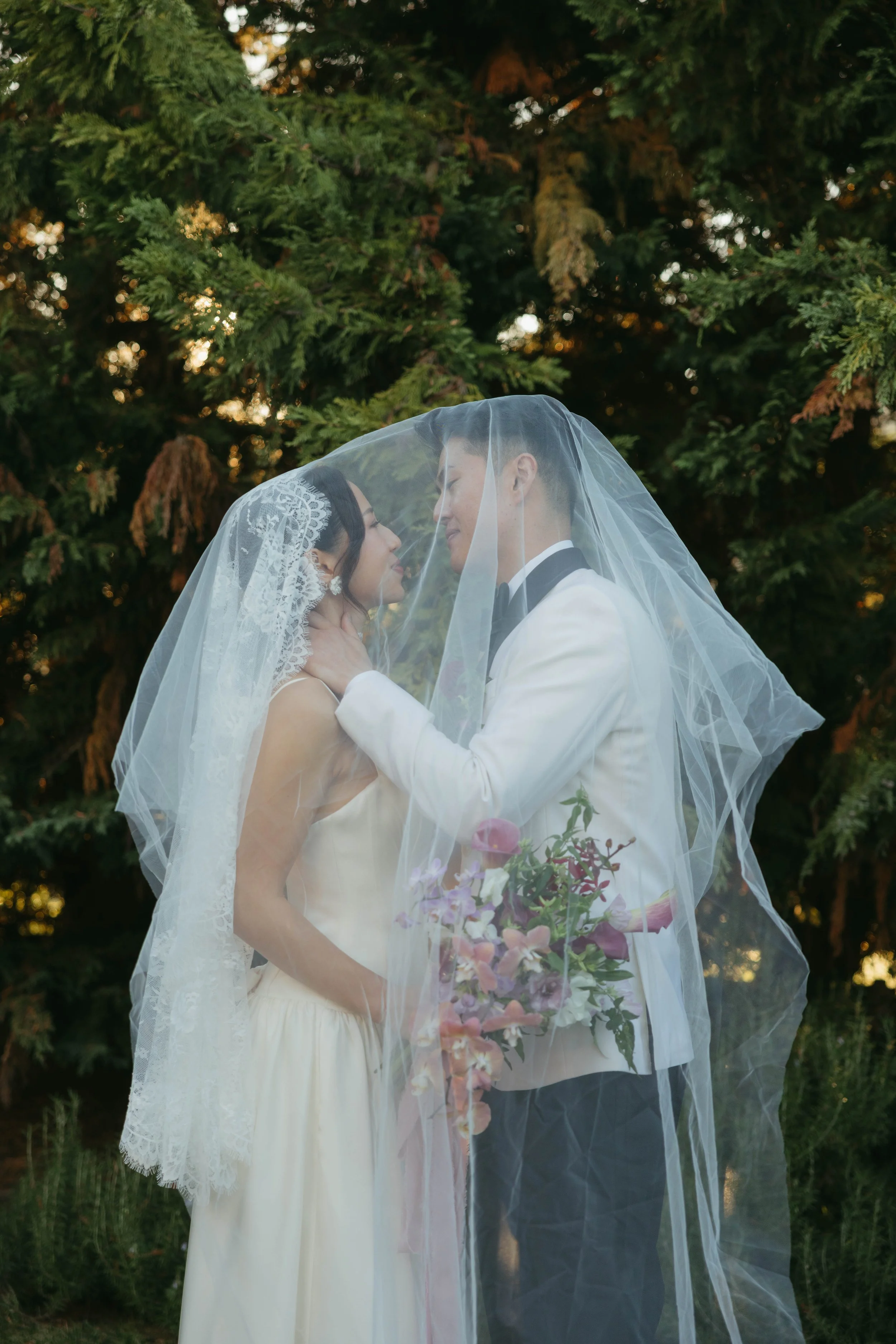 A bride and groom standing close together outdoors at sunset, under a sheer wedding veil, with greenery in the background. The bride holds a bouquet of flowers, and they are about to kiss.