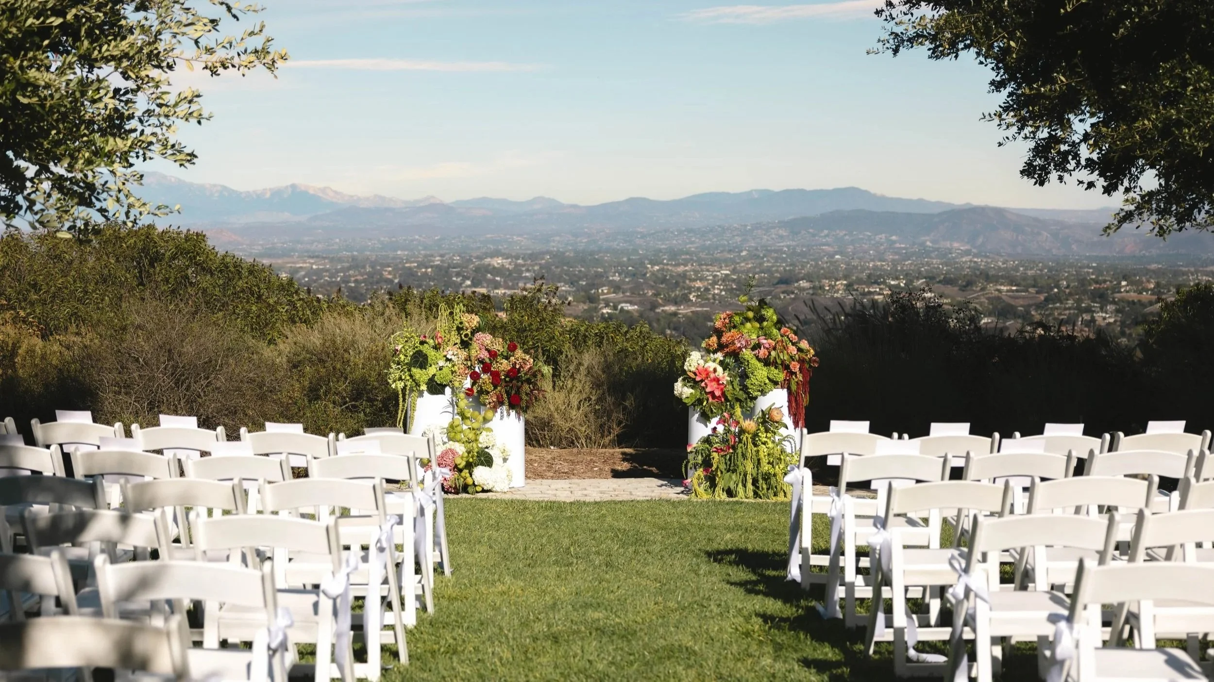 Outdoor wedding ceremony setup with white chairs and floral arrangements on white pedestals overlooking a scenic valley and distant mountains.