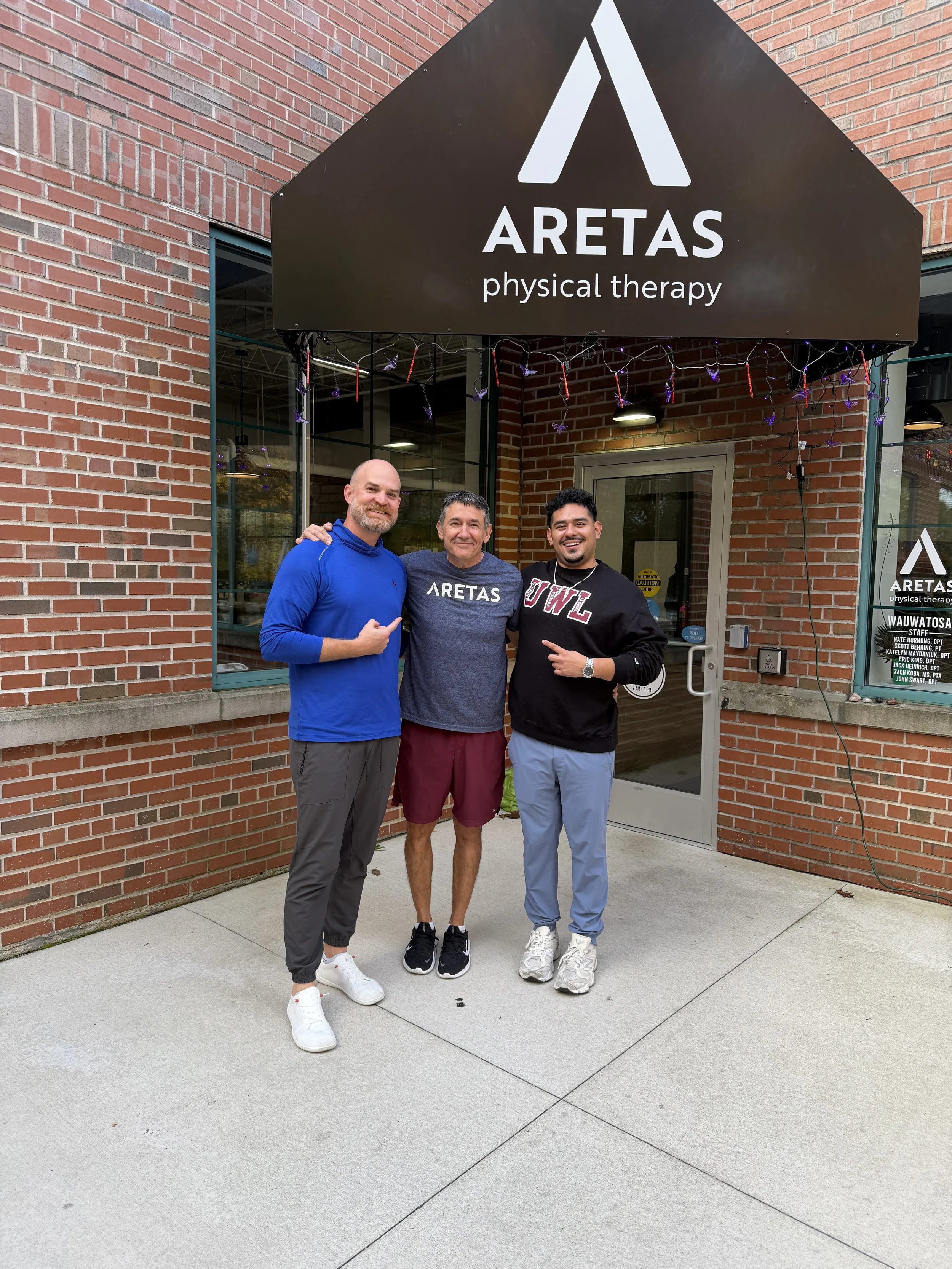Three men smiling and standing outside of a brick building with "Aretas Physical Therapy" written on the black awning