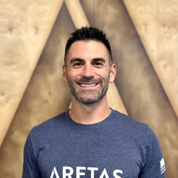 A smiling man with short dark hair and a beard, wearing a blue T-shirt with white text, standing in front of a textured wooden wall with geometric patterns.