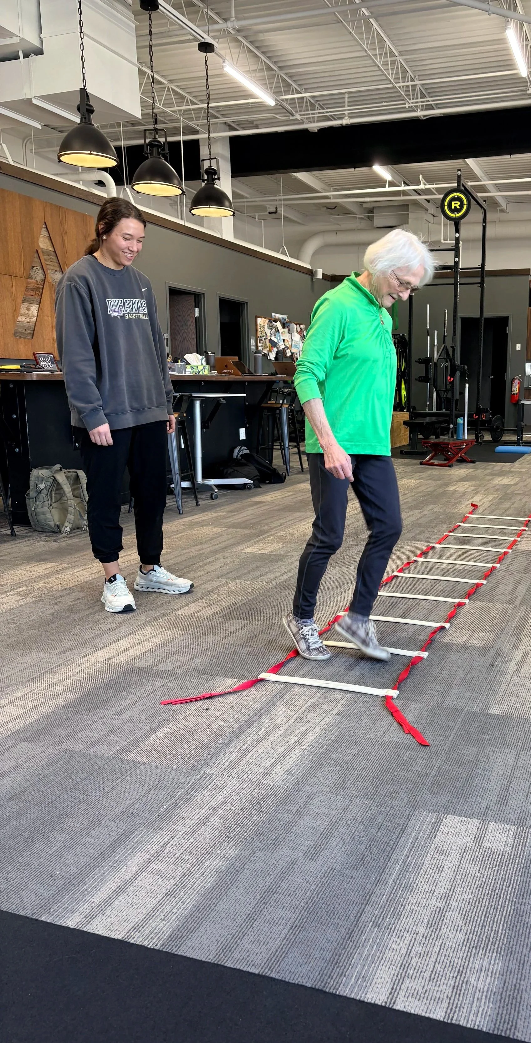 An older woman in a bright green shirt performs ladder agility drills, while a smiling, young female Aretas staff member observes form