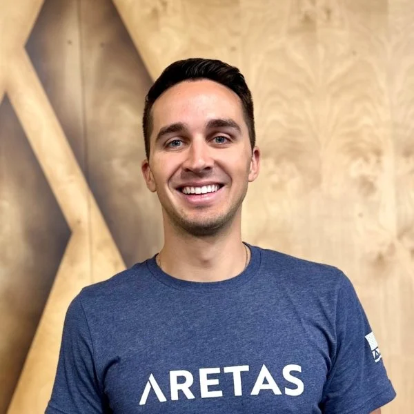 A smiling young man with short dark hair, wearing a blue T-shirt with the word 'ARETAS' printed on it, standing inside a room with wooden wall panels.