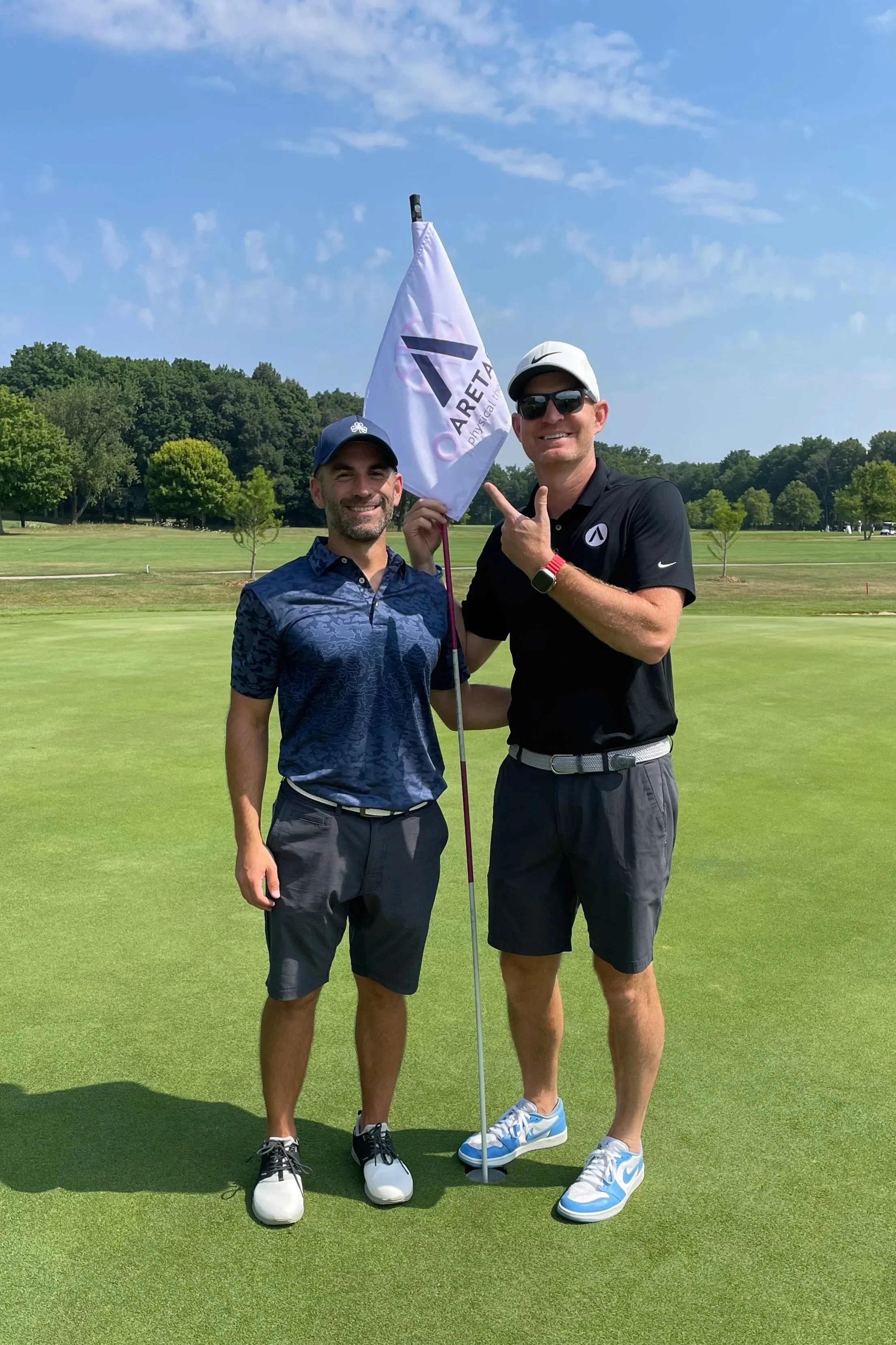 Nate and Kevin, Aretas owners, pose together and point at an Aretas golf flag during a golf tournament