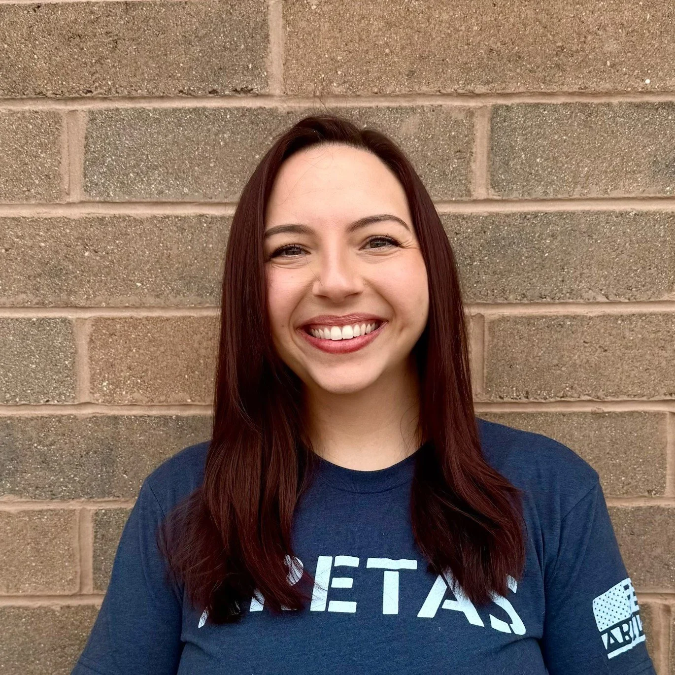 A young woman with shoulder-length reddish-brown hair and fair skin smiling, standing against a brick wall, wearing a navy blue t-shirt with white text and graphics.