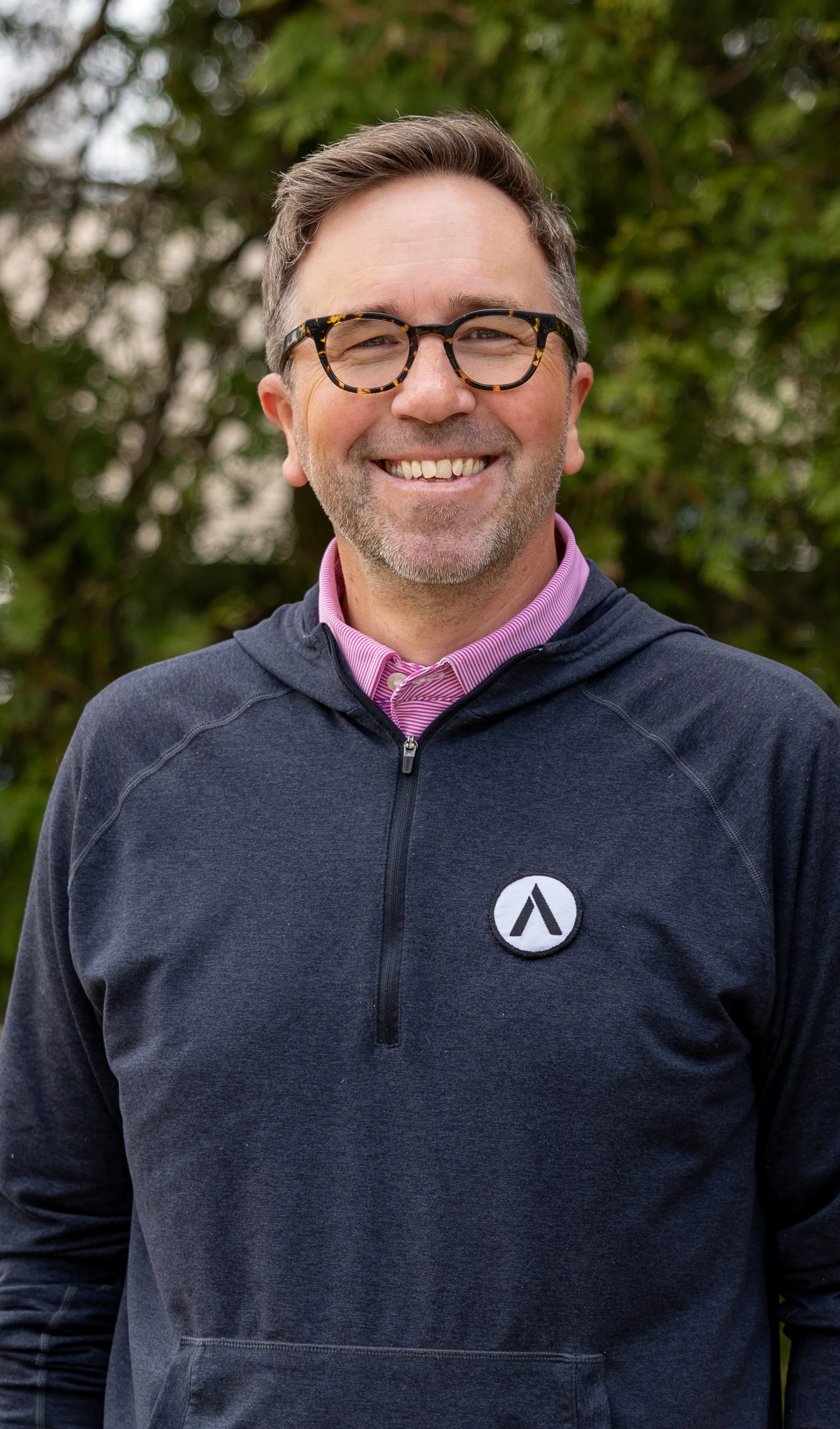 Smiling man wearing glasses, a pink collared shirt, and a black zip-up jacket with a logo on the chest, standing outdoors with green foliage in the background.