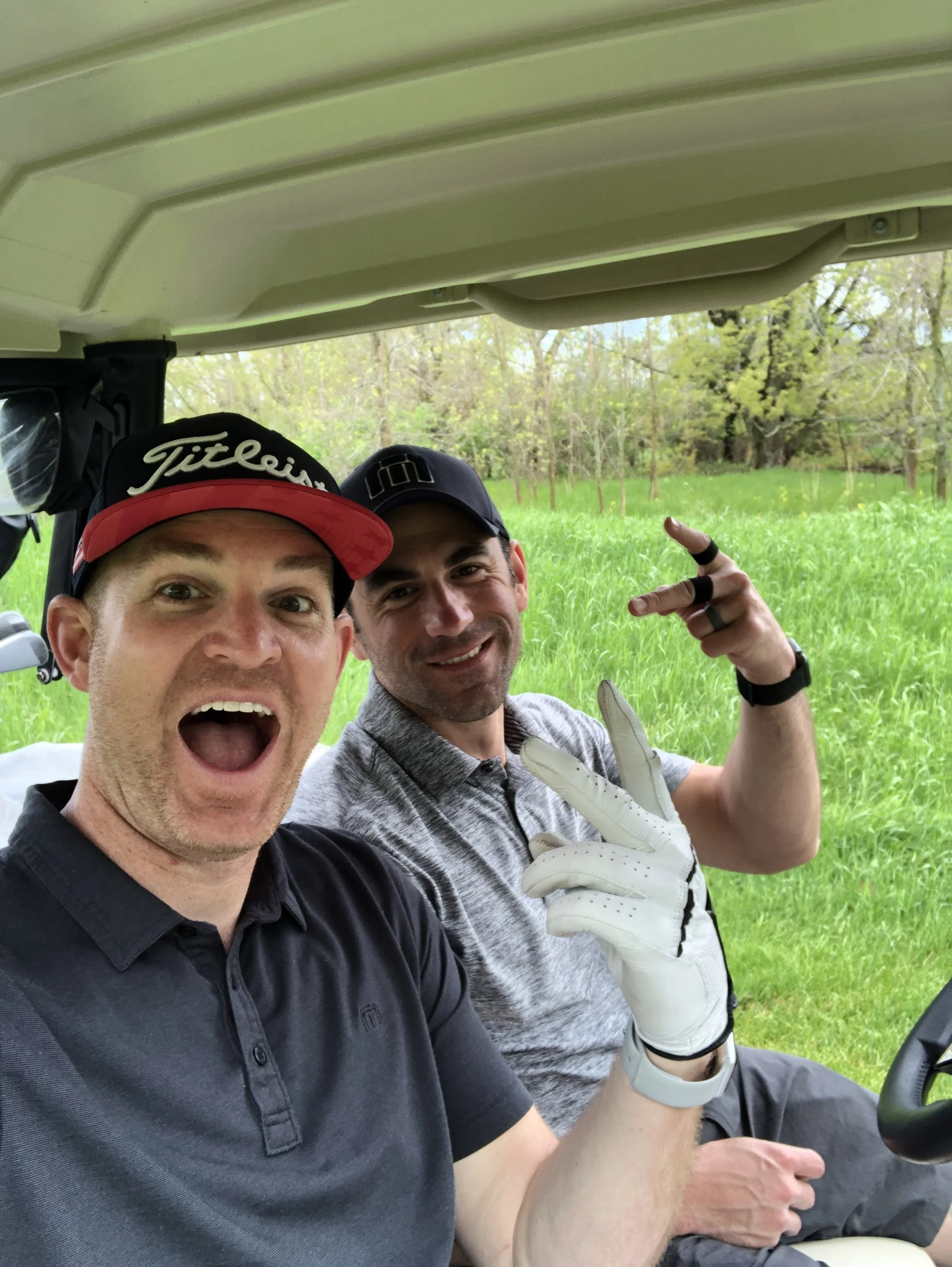 Two men sitting in a golf cart outdoors, smiling and making peace signs, with green grass and trees in the background.